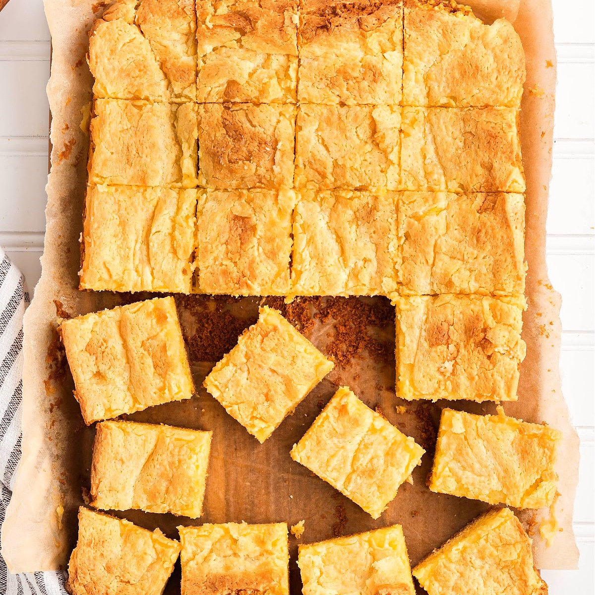 Gooey butter cake baked in a parchment-lined pan and cut into soft, golden squares with a crackled top.