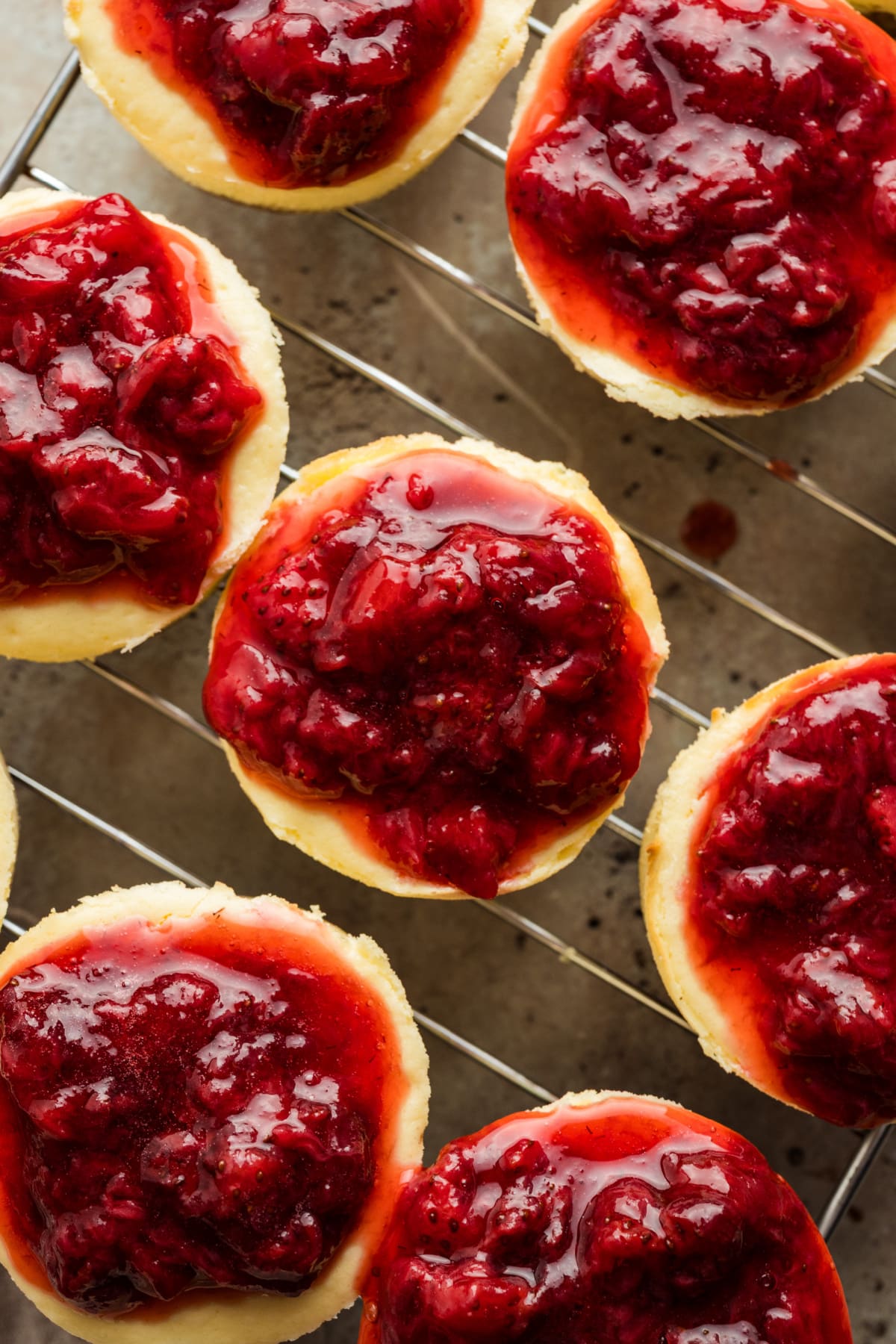 Baked mini cheesecakes with a thick strawberry topping set as they cool on a rack. Individual baked mini cheesecakes topped with a glossy strawberry topping, arranged closely together on a wire cooling rack.