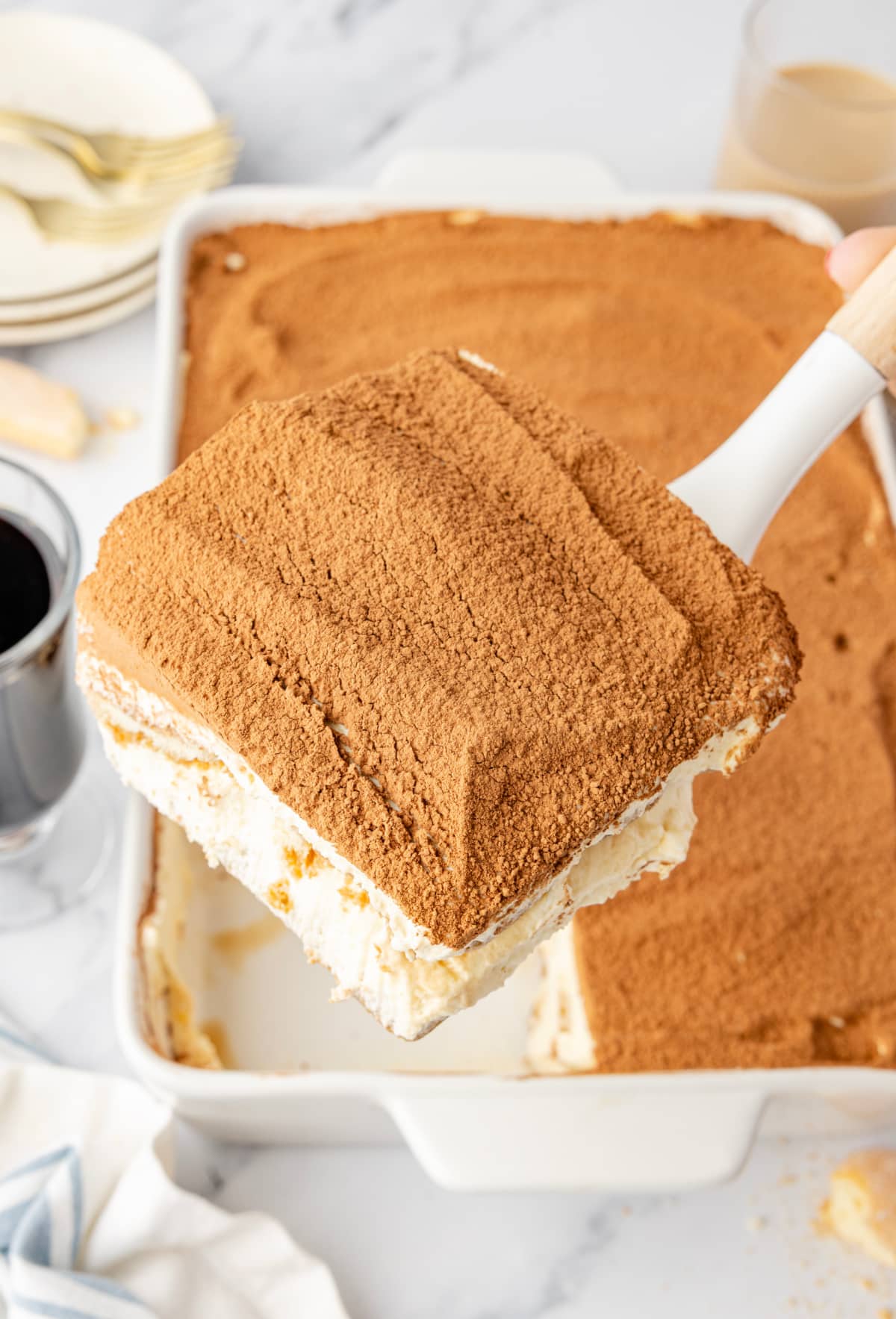 A serving spatula lifting a square of Baileys Irish Cream Tiramisu from a cocoa-dusted baking dish. Baileys Irish Cream Tiramisu being lifted from the dish, showing thick layers of mascarpone cream, espresso-soaked ladyfingers, and a cocoa-dusted top.