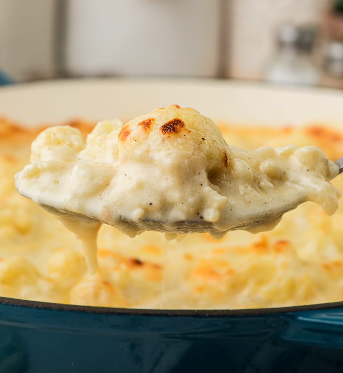 Spoonful of baked cauliflower coated in a thick, creamy cheese sauce, lifting from a skillet while still hot and melty.