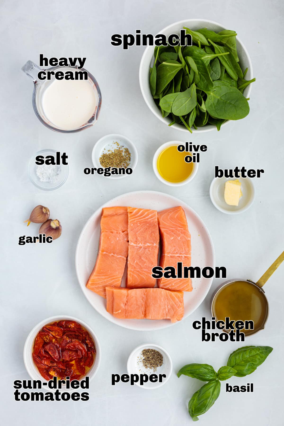 Measured ingredients for making creamy Tuscan salmon laid out before cooking. Ingredients for Tuscan salmon including salmon fillets, heavy cream, spinach, sun-dried tomatoes, garlic, olive oil, butter, chicken broth, oregano, salt, pepper, and fresh basil.