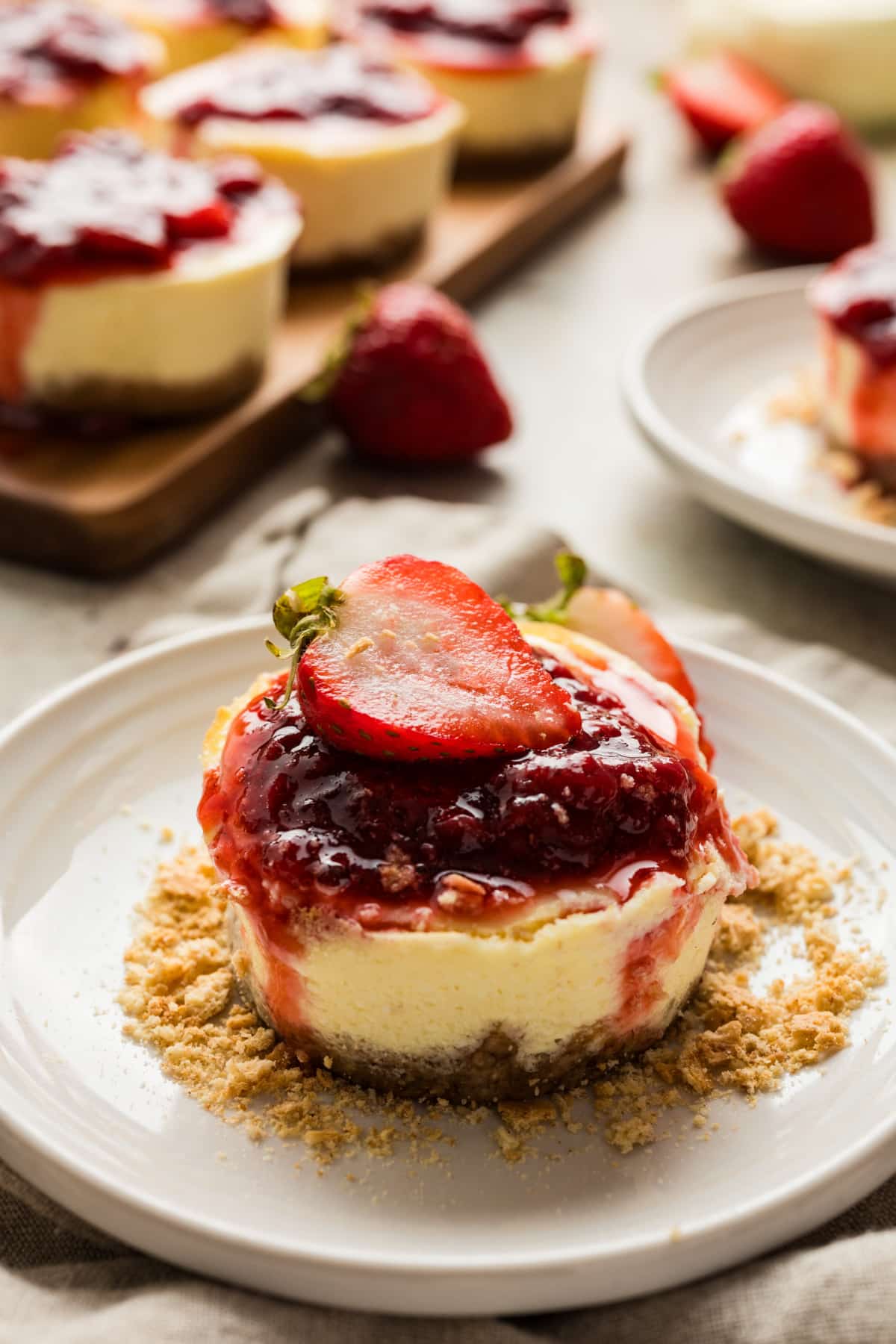 A plated strawberry mini cheesecake with a graham cracker crust and glossy strawberry topping. A single strawberry mini cheesecake topped with fresh strawberry sauce and a sliced strawberry, served on a white plate with graham cracker crumbs.