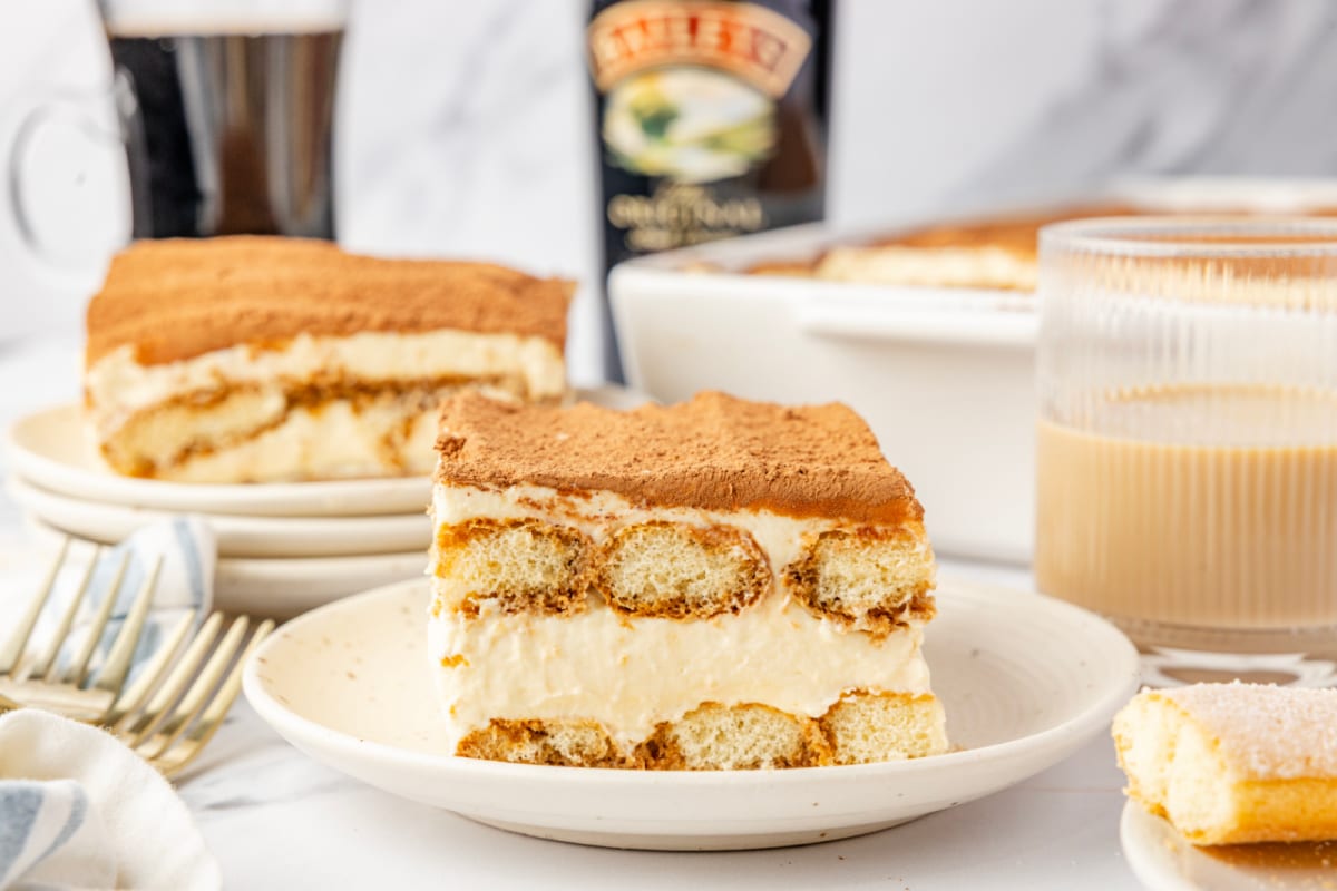 A plated slice of tiramisu showing espresso-soaked ladyfingers, mascarpone cream, and a cocoa-dusted top with Baileys Irish cream nearby. Slice of tiramisu on a plate with visible layers of espresso-soaked ladyfingers and mascarpone cream, with Baileys Irish cream and coffee in the background.