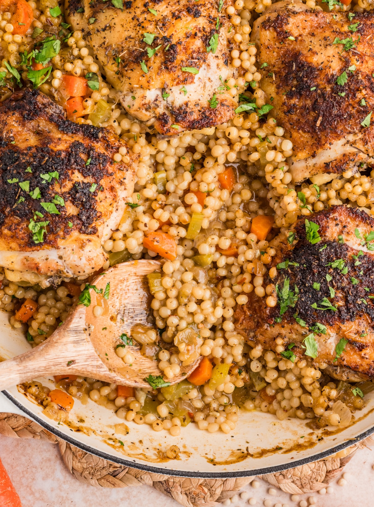Spoonful of pearl couscous with vegetables served alongside crispy baked chicken thighs in a wide pan.