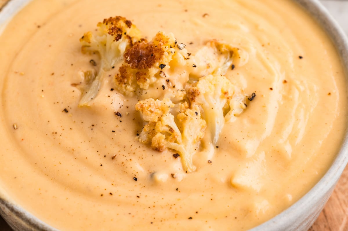 A close-up of creamy cauliflower soup topped with caramelized cauliflower florets and freshly cracked black pepper. Creamy cauliflower soup with roasted cauliflower florets and cracked black pepper blended smooth in a shallow bowl.