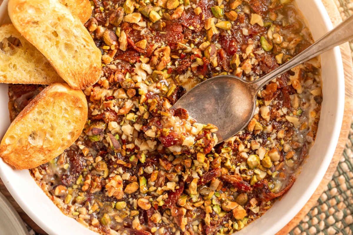 A spoon lifting a creamy scoop of baked Boursin dip layered with fig preserves, crisp bacon, and chopped pistachios and walnuts. A spoon scooping into a warm baked Boursin dip with bacon, fig preserves, pistachios, walnuts, and toasted crostini on the side.