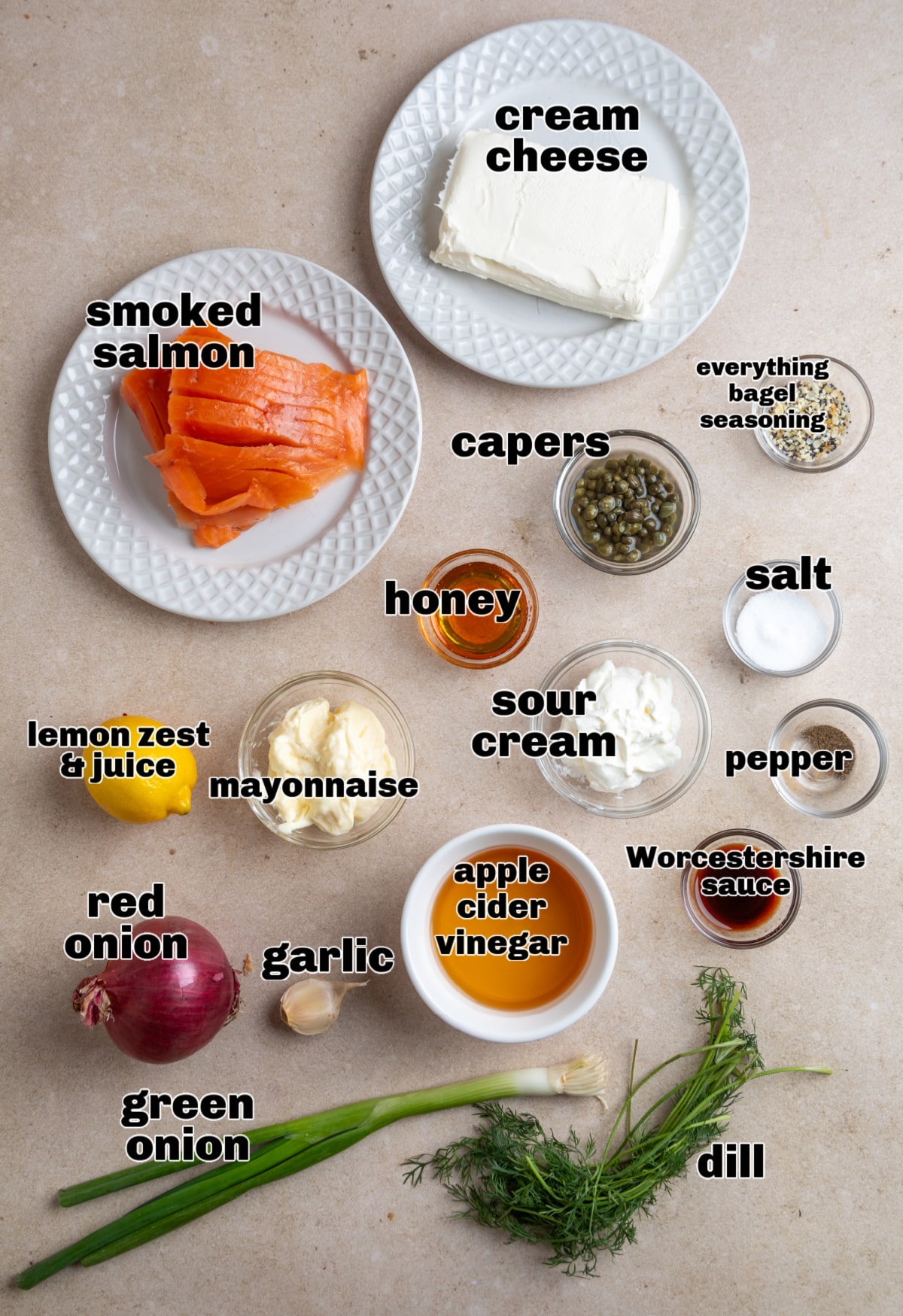 Ingredients for a smoked salmon cream cheese dip arranged on a light surface, including smoked salmon, cream cheese, sour cream, capers, dill, lemon, and red onion.