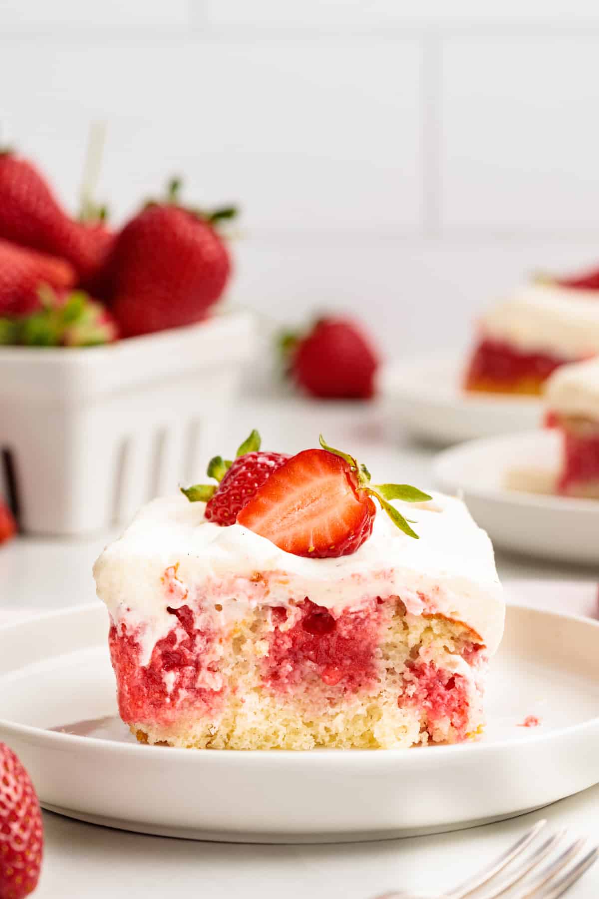 Strawberry poke cake slice with whipped vanilla cream cheese frosting and a fresh strawberry on top, showing the soaked strawberry filling throughout the cake.