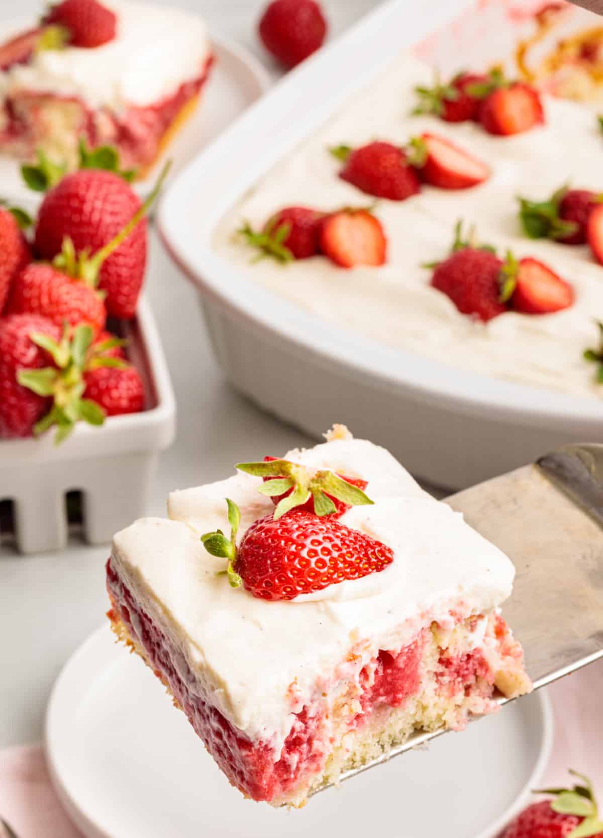 Slice of strawberry cake made from white cake mix with a soaked strawberry filling and whipped vanilla cream cheese frosting, lifted from the pan on a metal spatula.