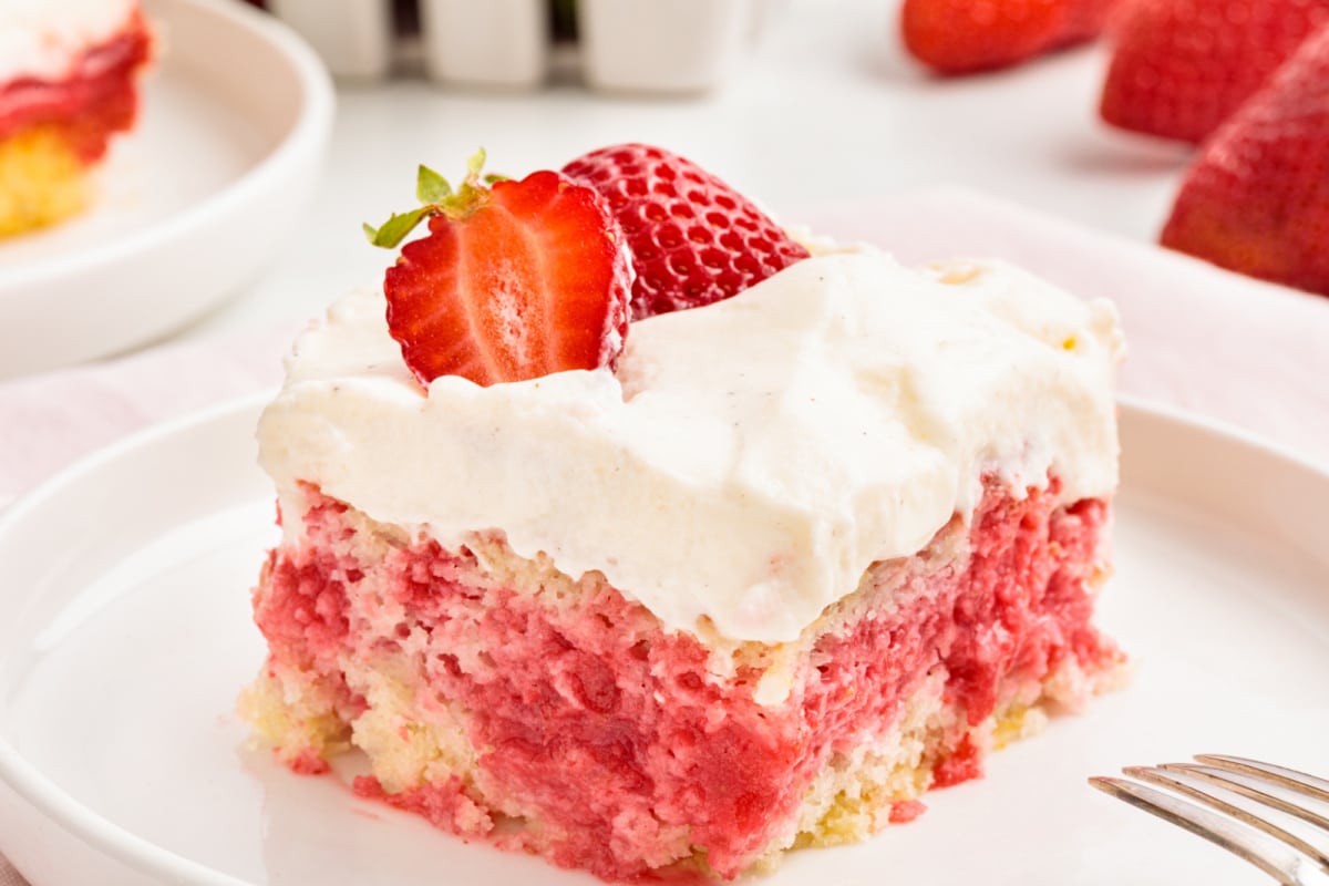 Strawberry poke cake slice with cream cheese frosting showing soaked strawberry filling layered through the cake and topped with a fresh strawberry.