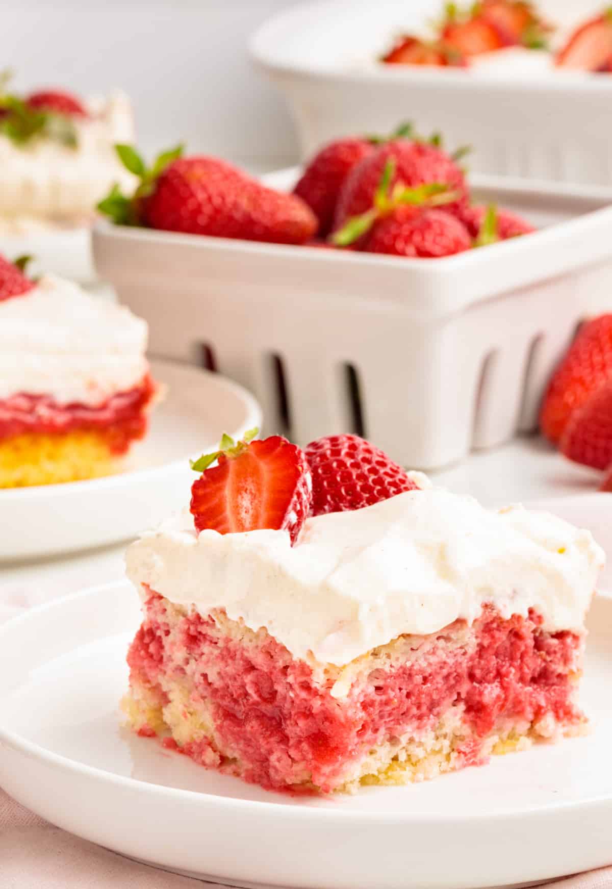 Strawberry poke cake slice showing pink strawberry filling soaked into white cake and topped with whipped vanilla cream cheese frosting and fresh strawberries.