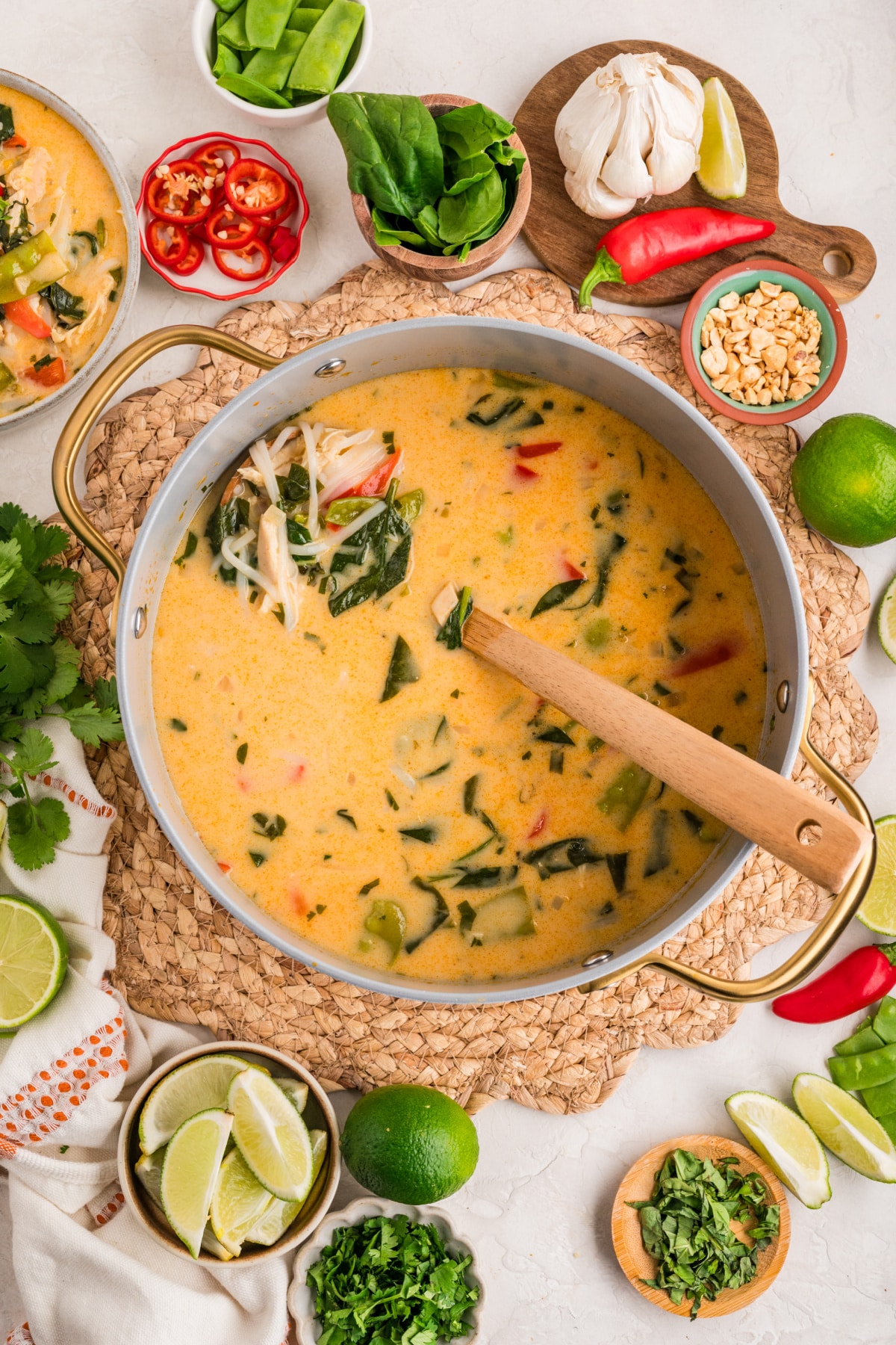 Thai chicken curry soup simmering in a wide pan with coconut milk, shredded chicken, snow peas, spinach, and herbs, with lime wedges and garnishes nearby.