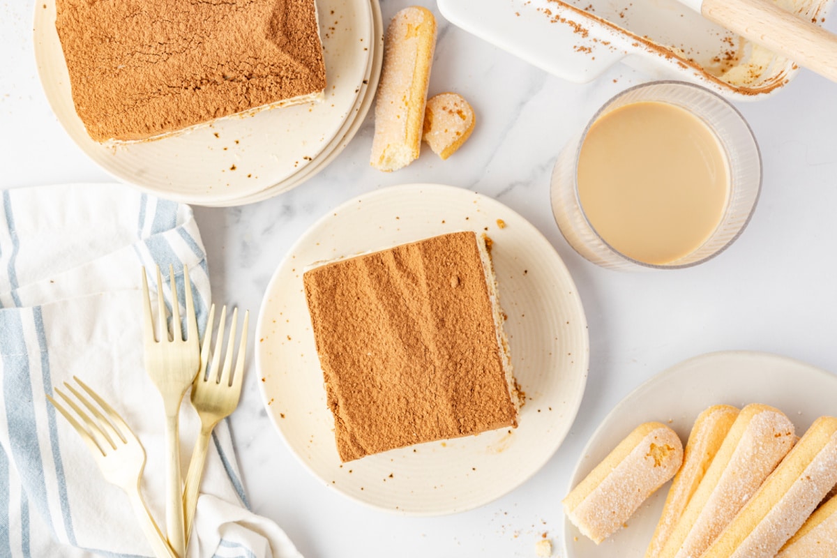 An overhead look at plated tiramisu with cocoa-dusted tops, ladyfingers, and a small glass of Irish cream alongside. Overhead view of plated tiramisu squares dusted with cocoa powder, with ladyfingers and a glass of Irish cream coffee on the side.