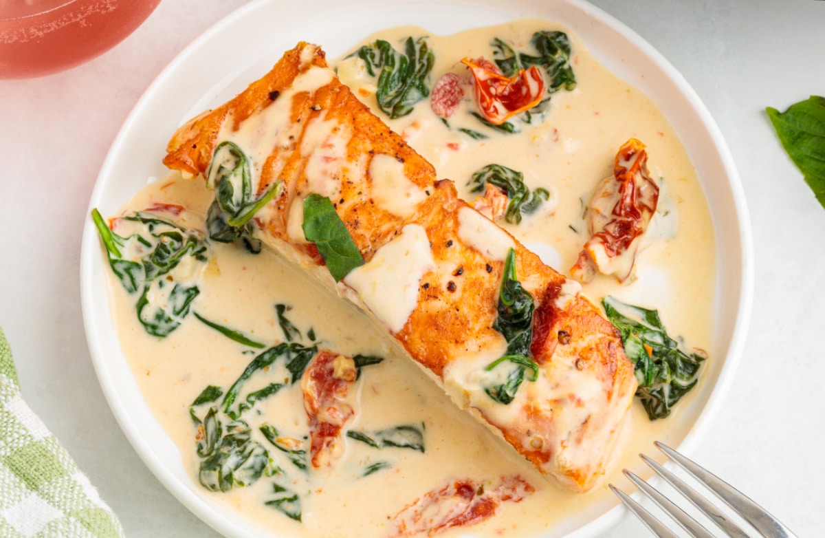 A plated portion of creamy Tuscan salmon finished with sun-dried tomatoes and spinach in a garlic cream sauce. Creamy Tuscan salmon served in a shallow bowl with garlic cream sauce, sun-dried tomatoes, and wilted spinach.