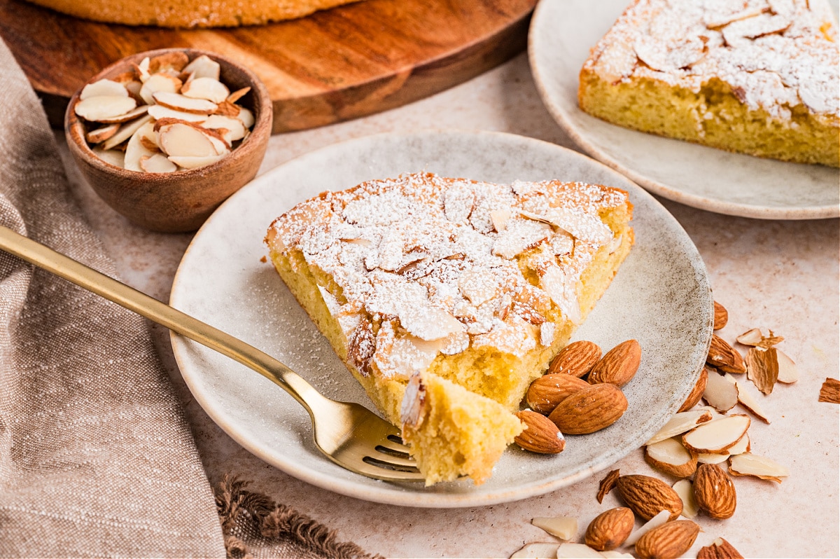 Close view of a slice of almond cake with a bite removed, highlighting the tender crumb and powdered sugar topping. Almond cake slice with a forkful taken out, showing the soft interior and sliced almonds on top