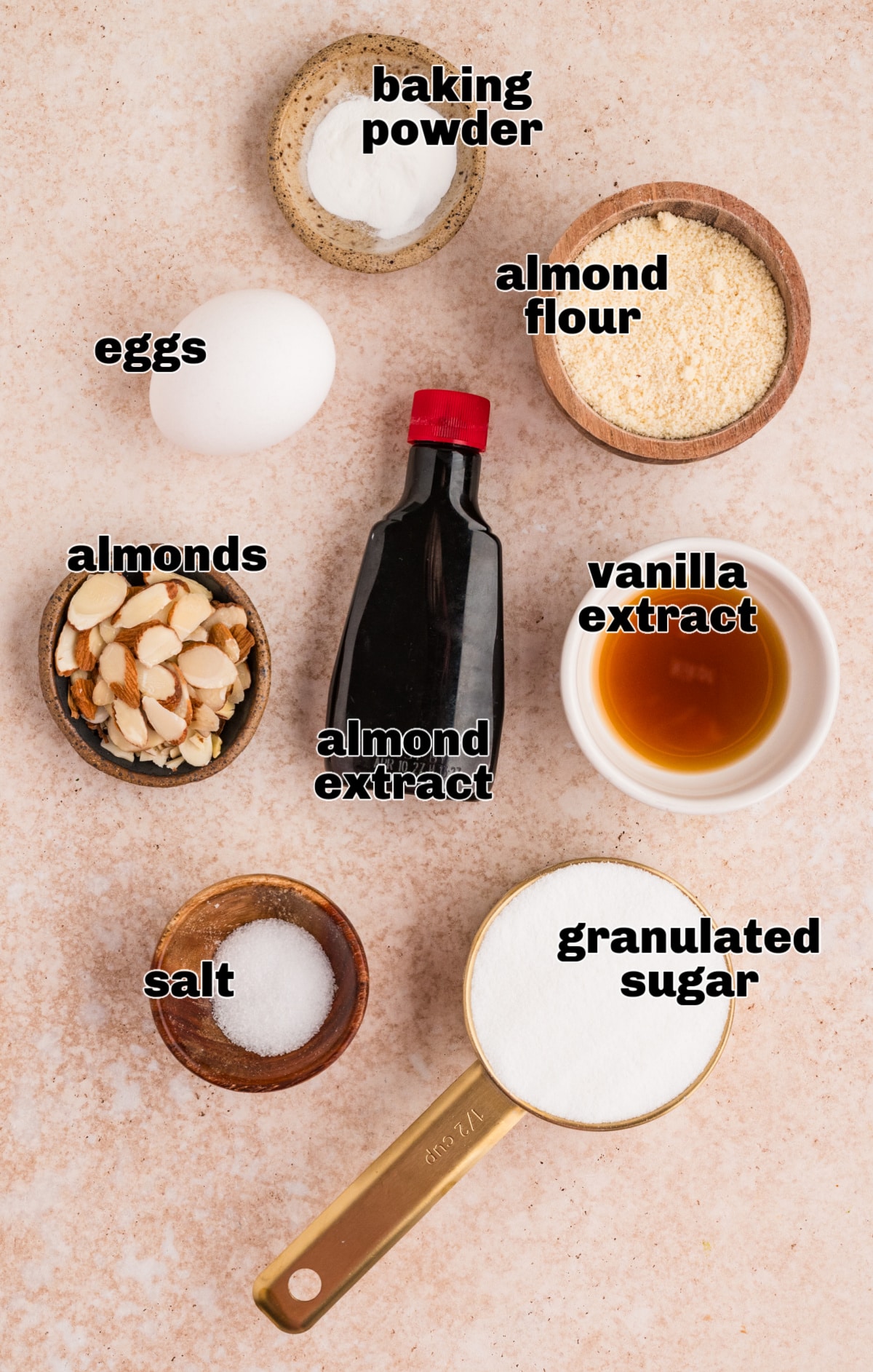Overhead view of measured ingredients arranged on a neutral surface for making almond cake. Ingredients for almond cake including almond flour, eggs, sugar, baking powder, sliced almonds, vanilla extract, almond extract, and salt