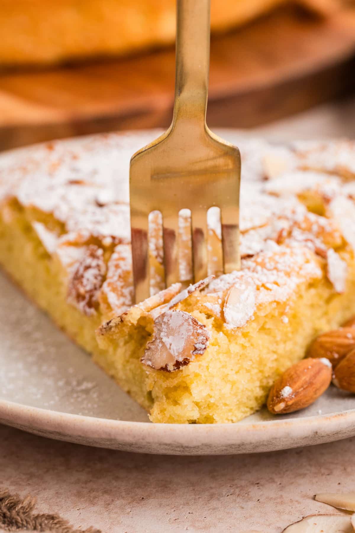 Tight detail of a tender almond flour cake slice topped with powdered sugar and sliced almonds, showing the moist interior texture. Close-up of almond flour cake slice with fork pressing into the soft crumb and sliced almonds on top
