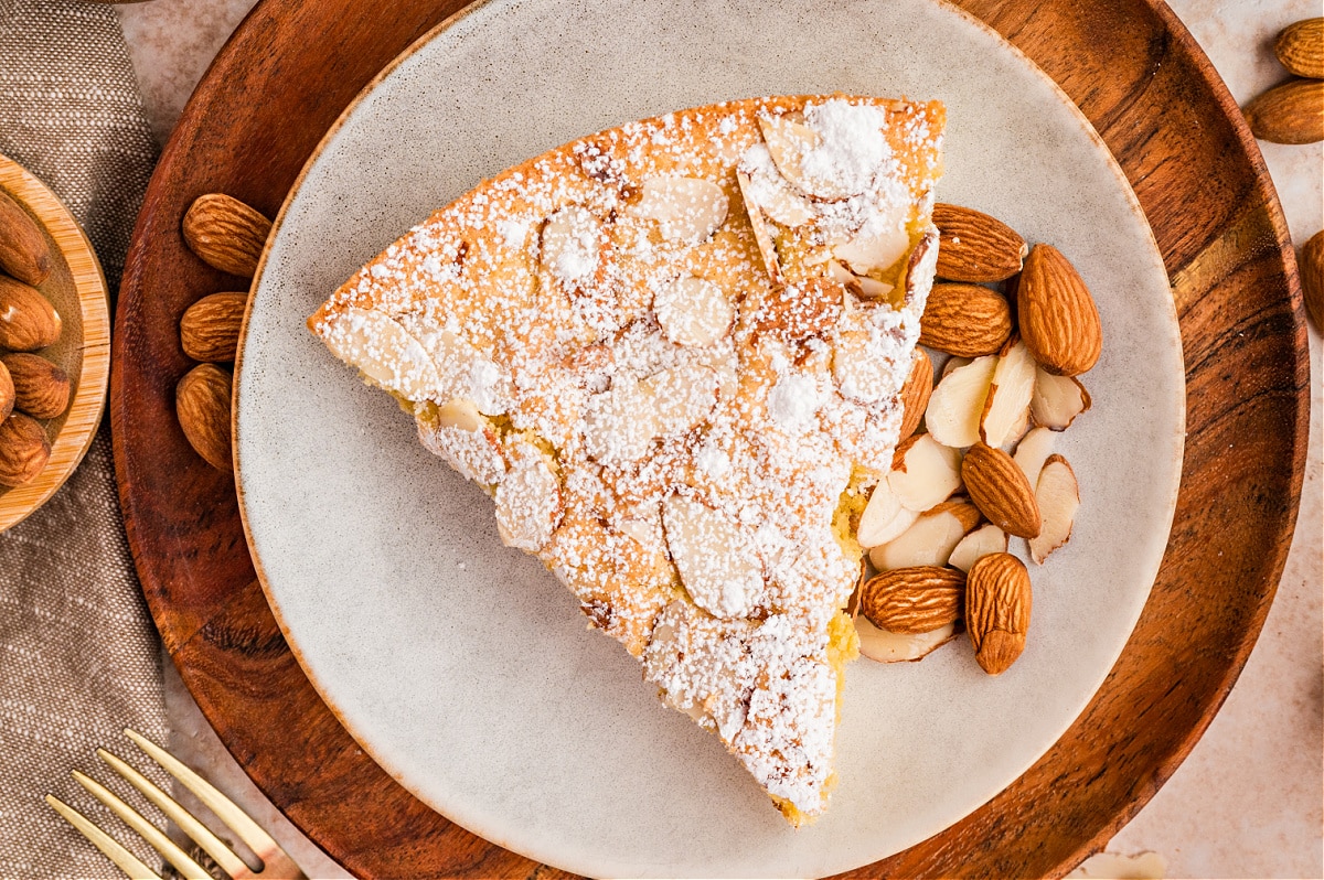 Top view of a single slice of almond cake served on a neutral plate with whole and sliced almonds scattered around. Overhead view of almond cake slice dusted with powdered sugar and topped with sliced almonds on a plate