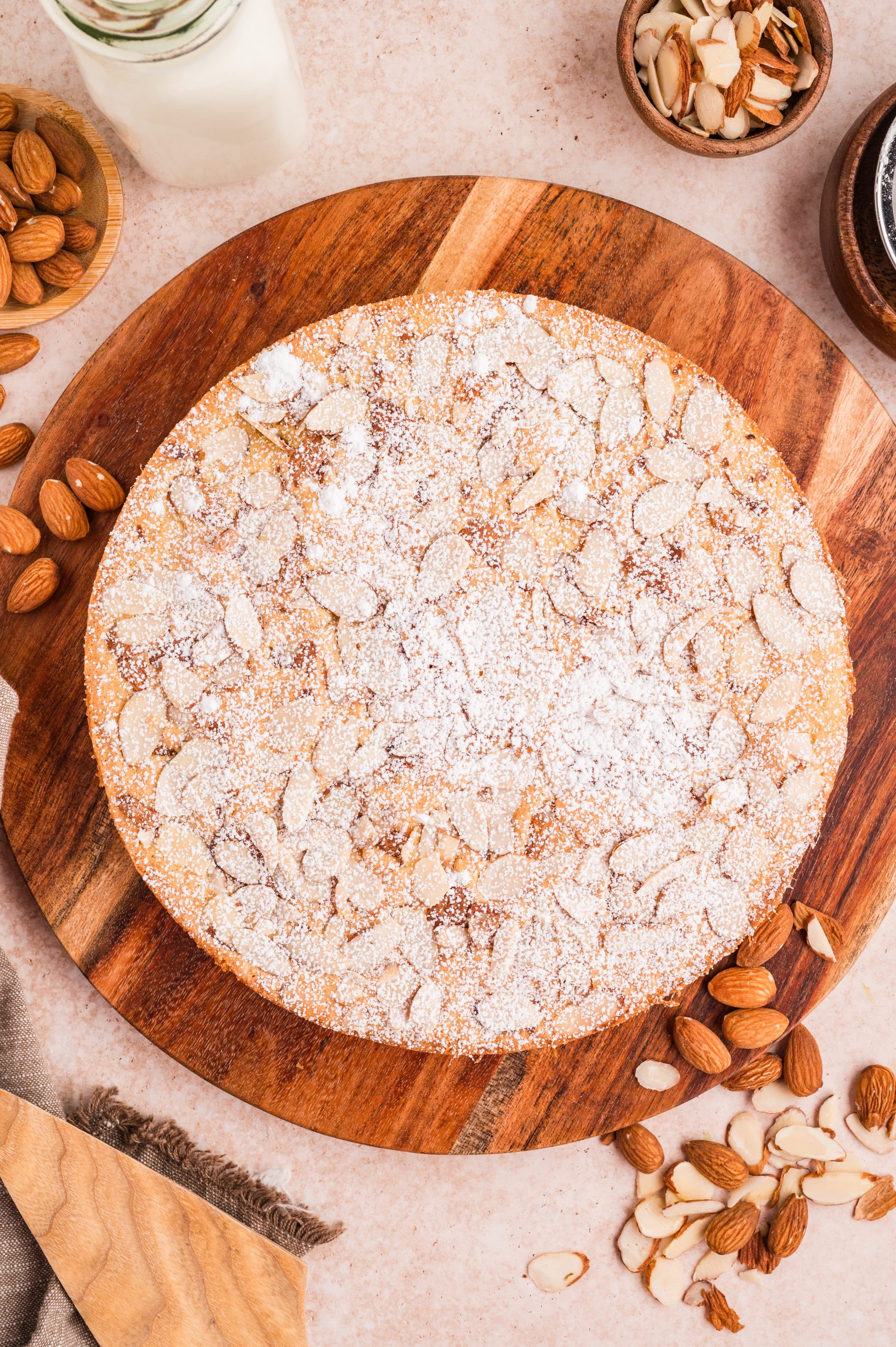 Overhead view of a whole round almond flour cake finished with sliced almonds and powdered sugar before slicing. Whole almond flour cake topped with sliced almonds and a dusting of powdered sugar on a wooden board
