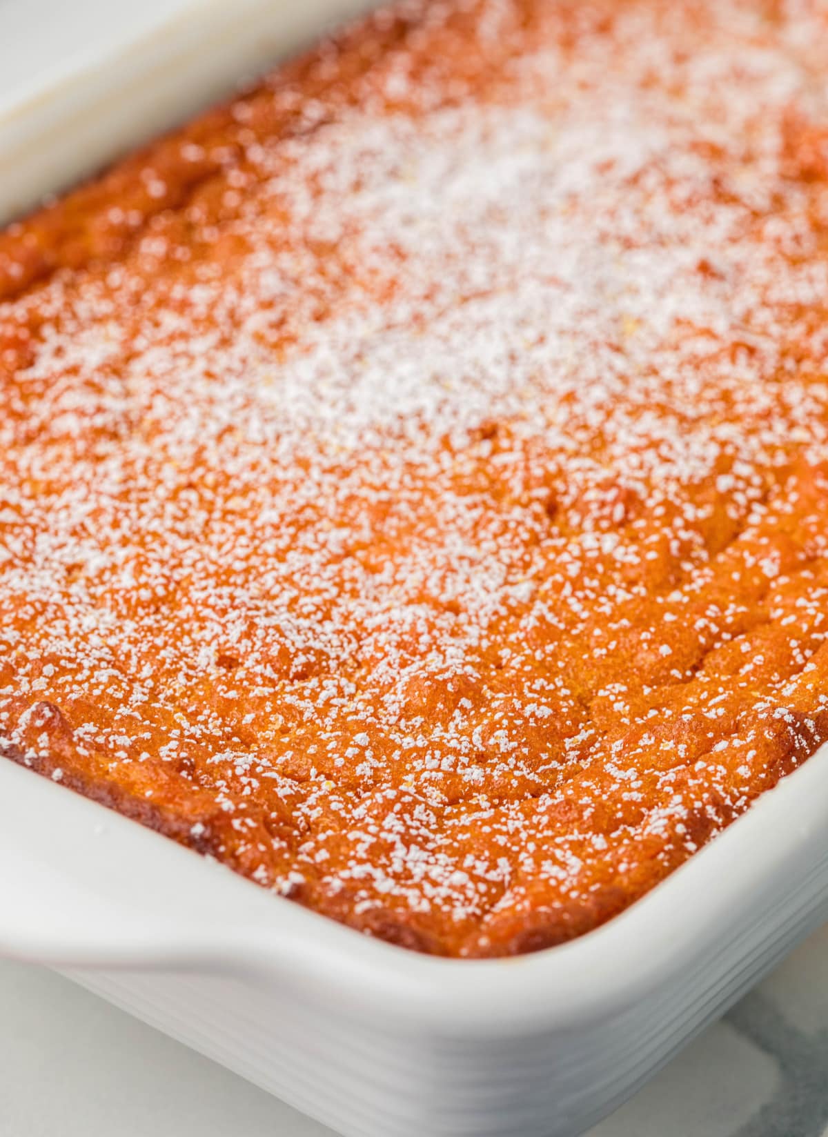 Sweet carrot casserole baked in white dish Powdered sugar being dusted over freshly baked Carrot Souffle in an 11x7 baking dish