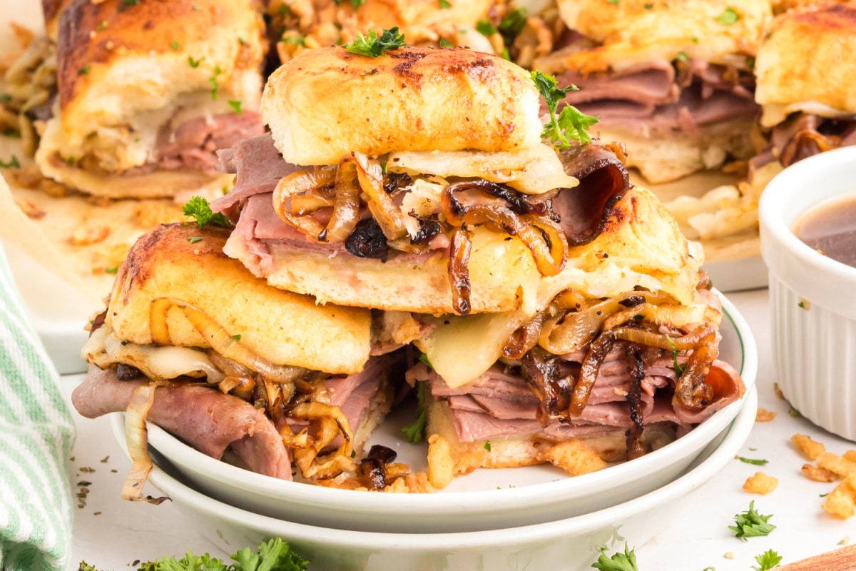 Close-up of baked French dip sliders filled with thin-sliced roast beef, provolone cheese, caramelized onions, and served with au jus for dipping French dip sliders stacked with roast beef, melted provolone, caramelized onions, and au jus