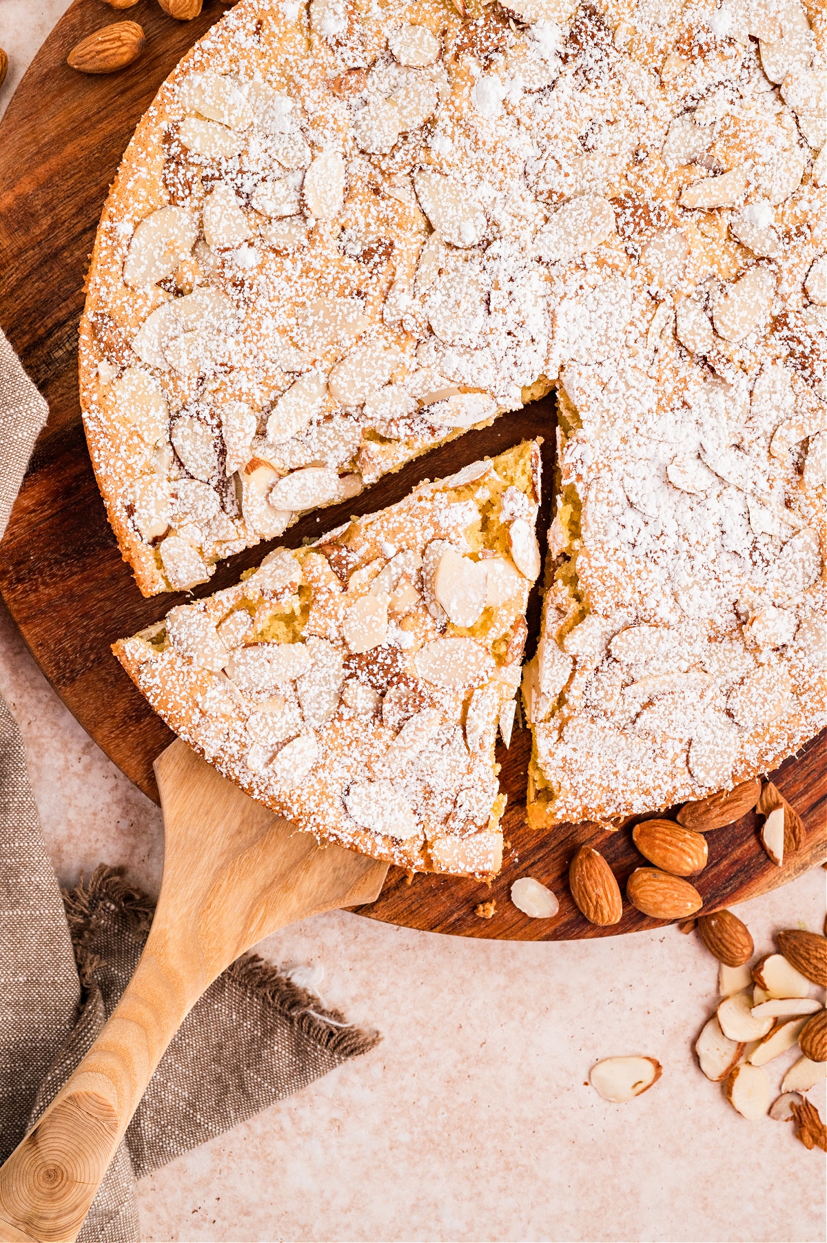 Top view of a round almond flour cake with one slice removed, showing the tender interior and almond topping. Overhead view of sliced almond flour cake with powdered sugar and toasted sliced almonds