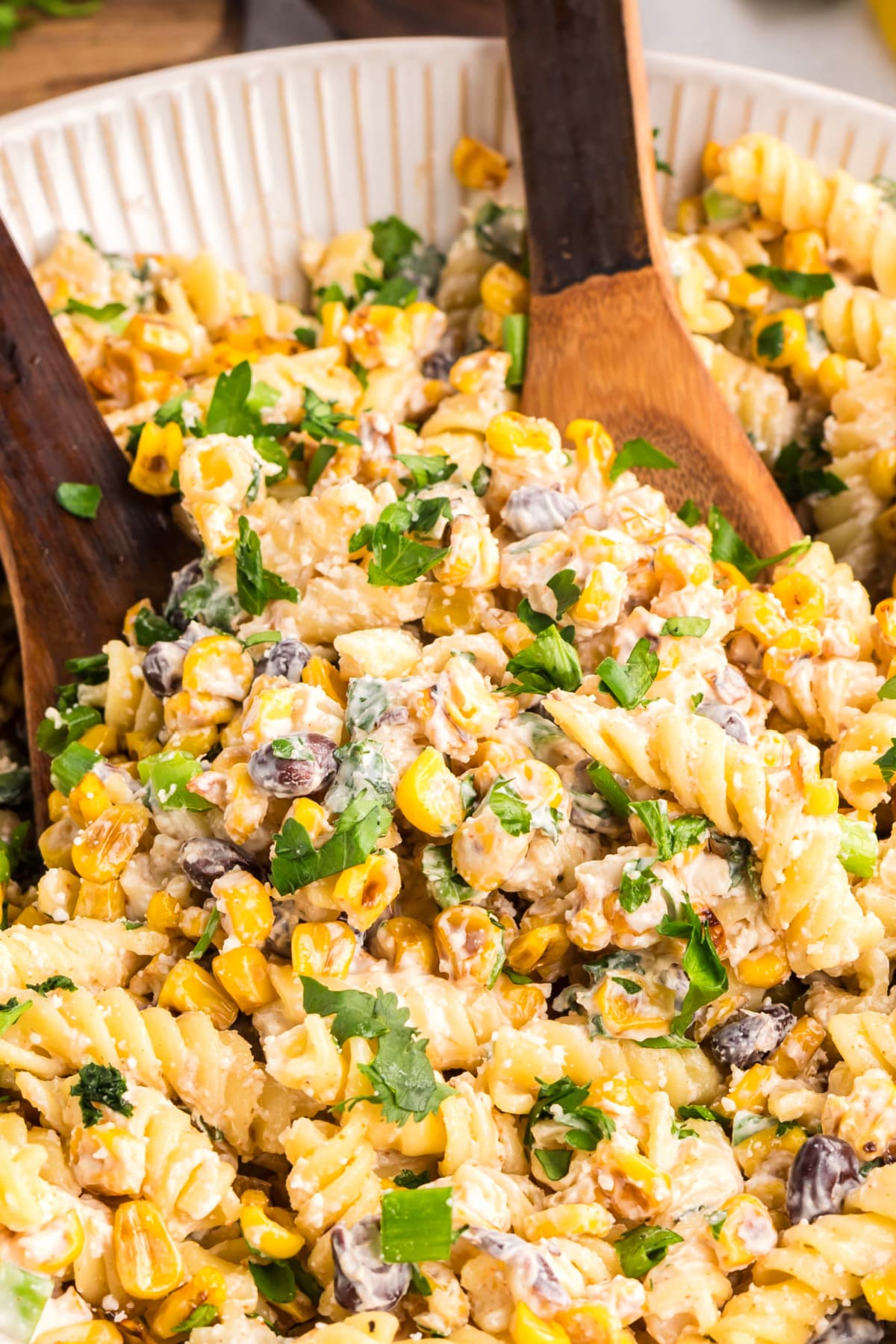 Close-up of creamy corn pasta salad being served with wooden spoons Creamy elote-style pasta salad with roasted corn, rotini, black beans, and fresh herbs