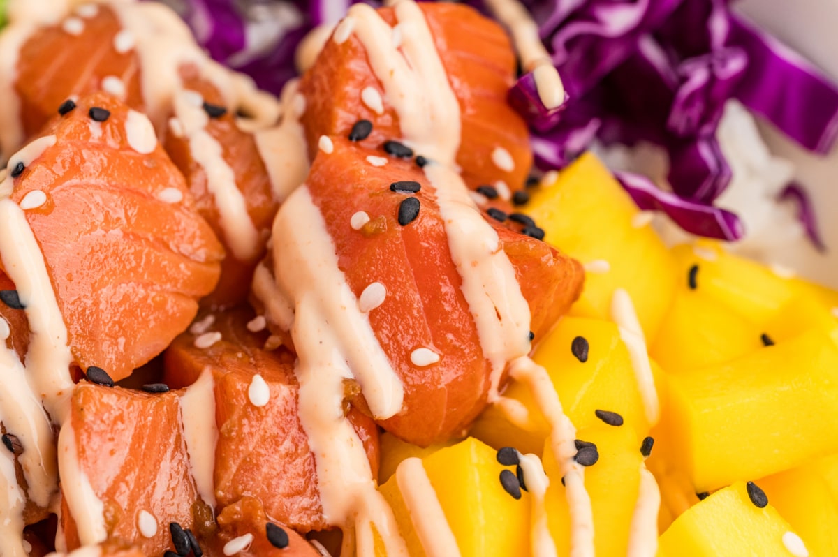 Close-up of marinated salmon cubes drizzled with sauce beside fresh mango and shredded purple cabbage Salmon poke bowls with marinated raw salmon, mango, sesame seeds, and sriracha mayo
