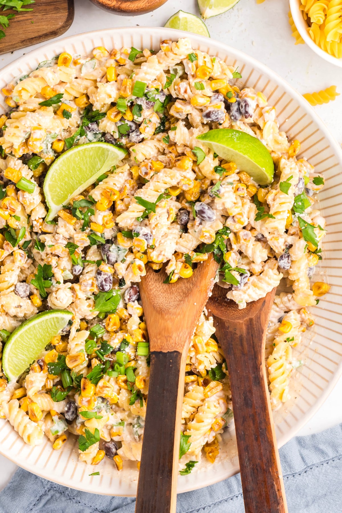 Creamy pasta salad tossed with corn, herbs, and lime, shown with serving spoons Mexican street corn pasta salad with rotini, roasted corn, black beans, cotija, and lime wedges