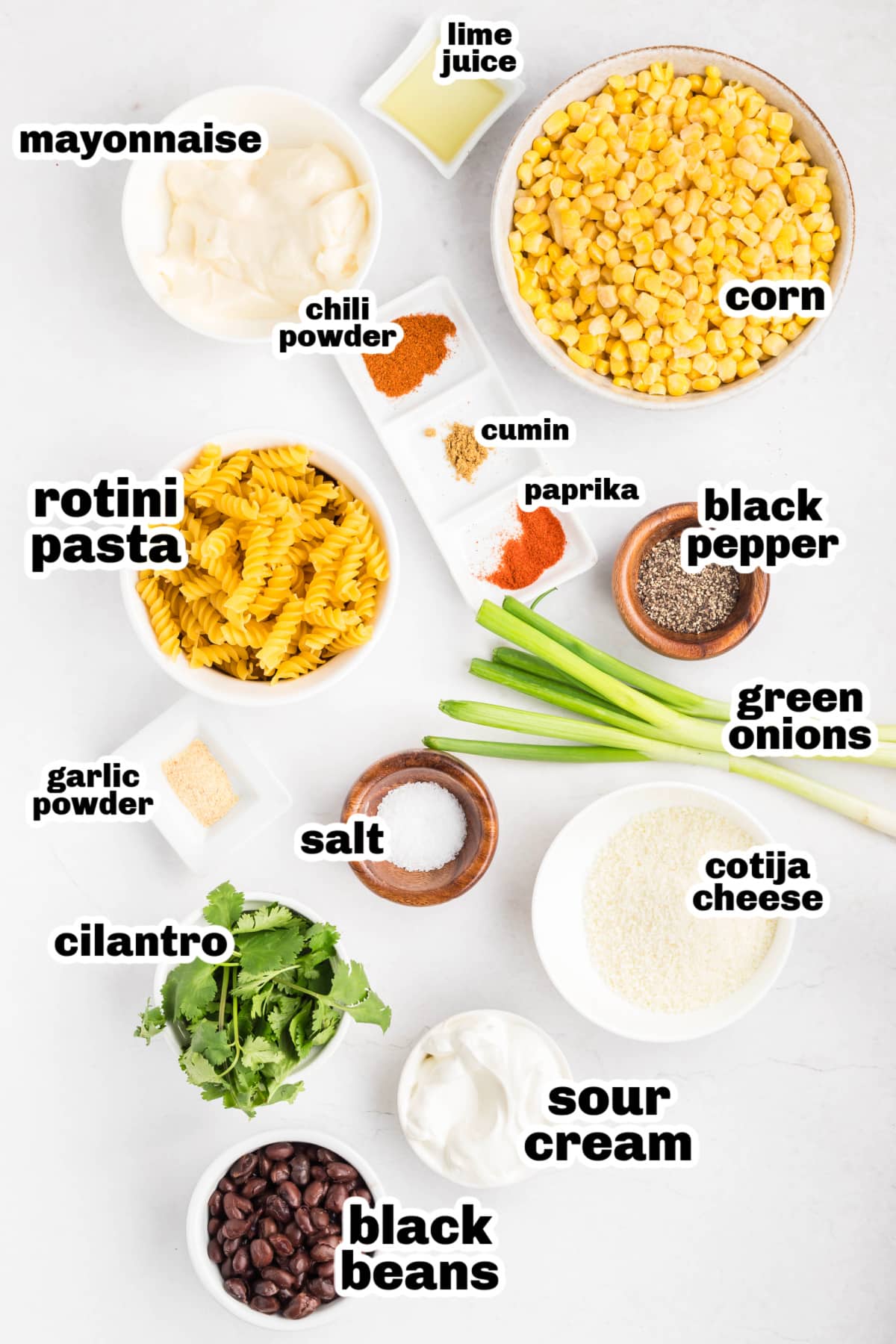 Overhead view of measured ingredients for a creamy street corn pasta salad, featuring rotini pasta, corn kernels, cotija cheese, cilantro, green onions, black beans, sour cream, mayonnaise, lime juice, chili powder, garlic powder, cumin, paprika, salt, and black pepper Ingredients for street corn pasta salad including rotini pasta, corn, cotija cheese, cilantro, green onions, black beans, sour cream, mayonnaise, lime juice, and spices