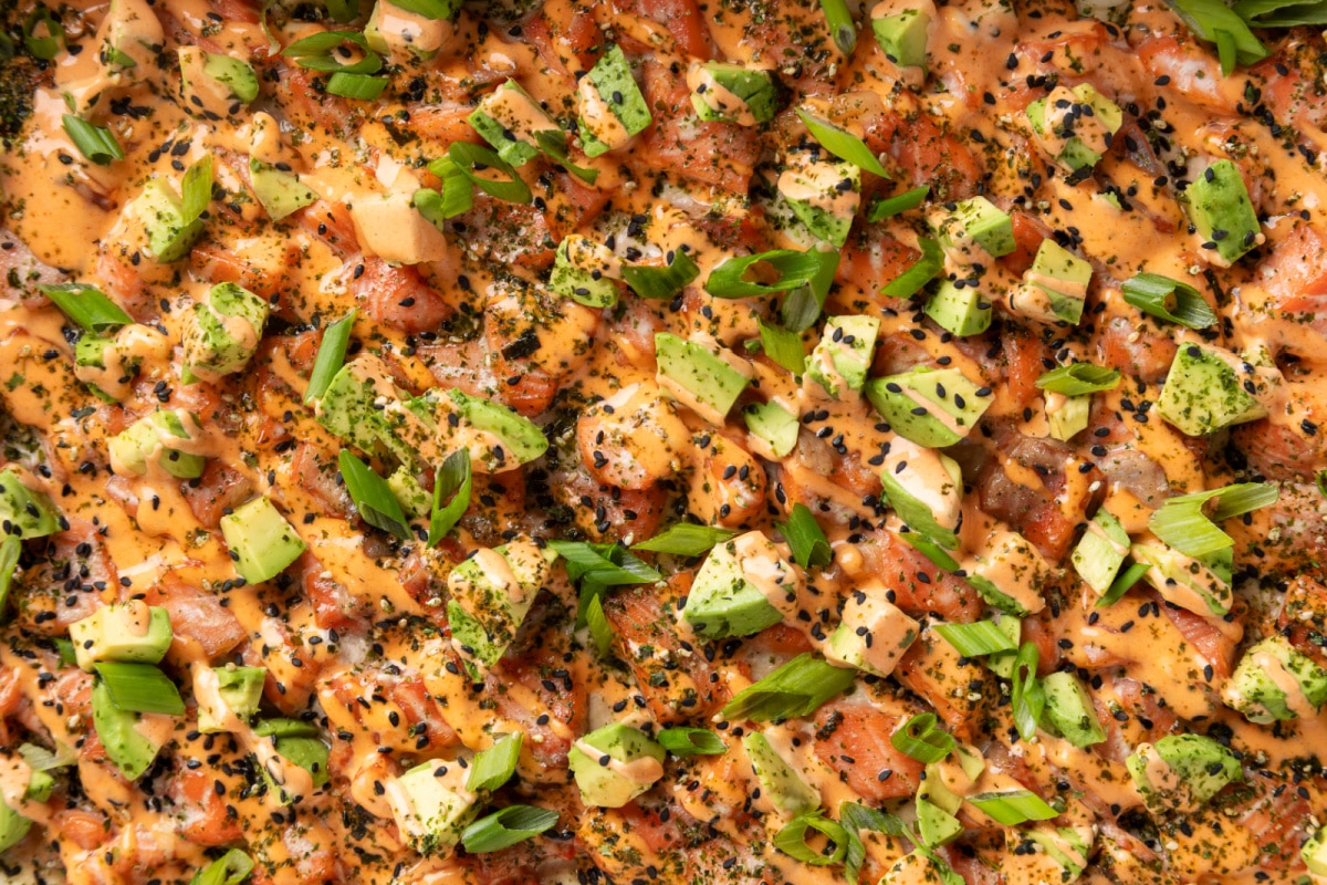 Close-up of Salmon Crab Sushi Bake showing creamy crab layer, baked salmon, and spicy mayo drizzle. Close-up of Salmon Crab Sushi Bake topped with spicy mayo, avocado, green onions, and black sesame seeds