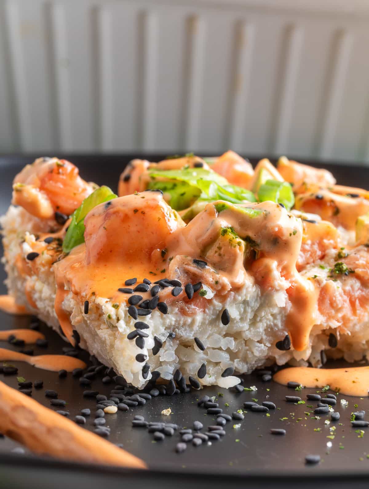 Salmon Crab Sushi Bake served in thick layered squares with creamy topping. Salmon Crab Sushi Bake slice with spicy mayo, sesame seeds, and green onions on a dark plate
