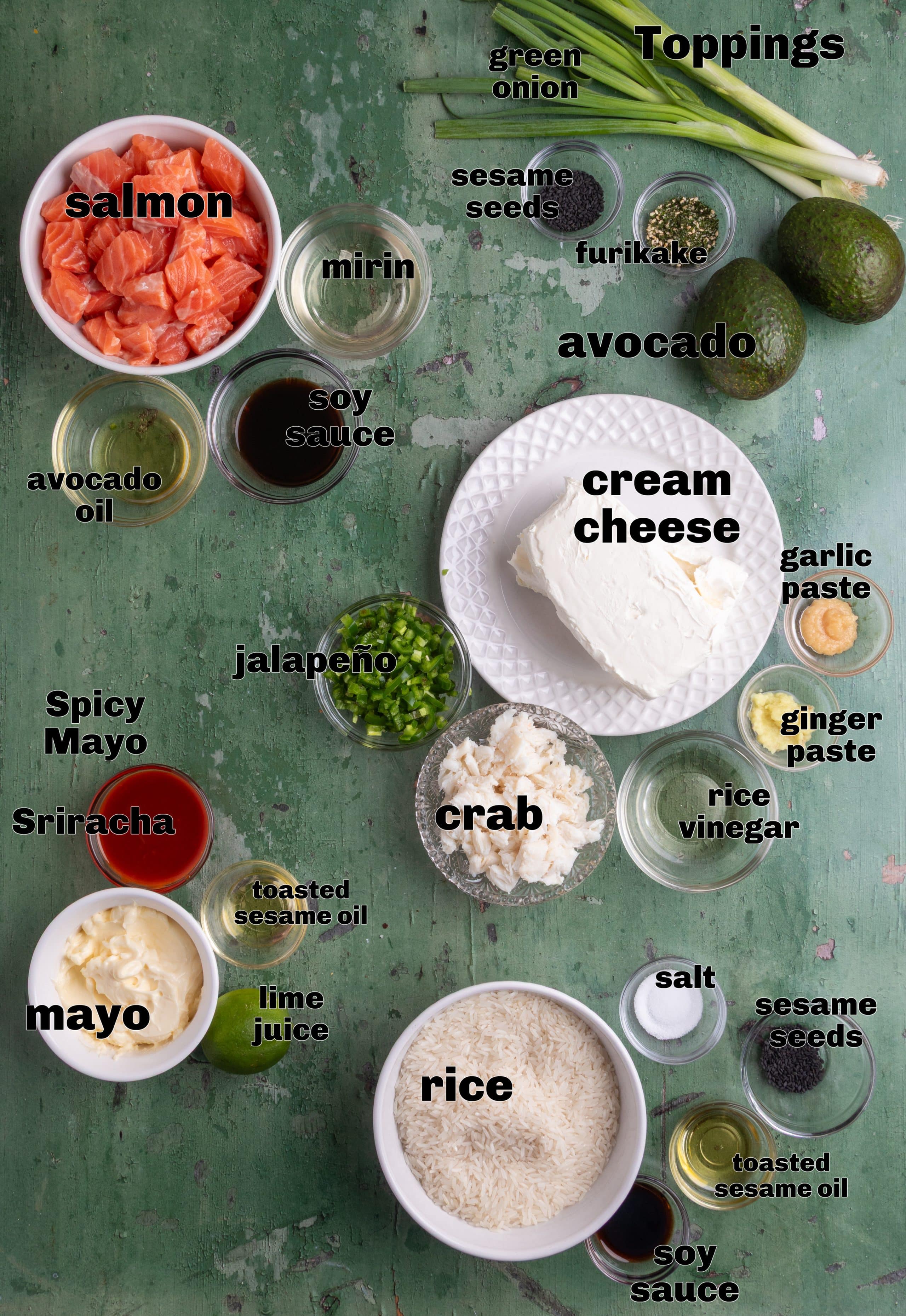 All ingredients needed for a layered sushi bake recipe arranged and labeled. Ingredients for sushi bake including salmon, lump crab, jasmine rice, cream cheese, jalapeño, soy sauce, mirin, mayonnaise, Sriracha, avocado, sesame seeds, and green onions
