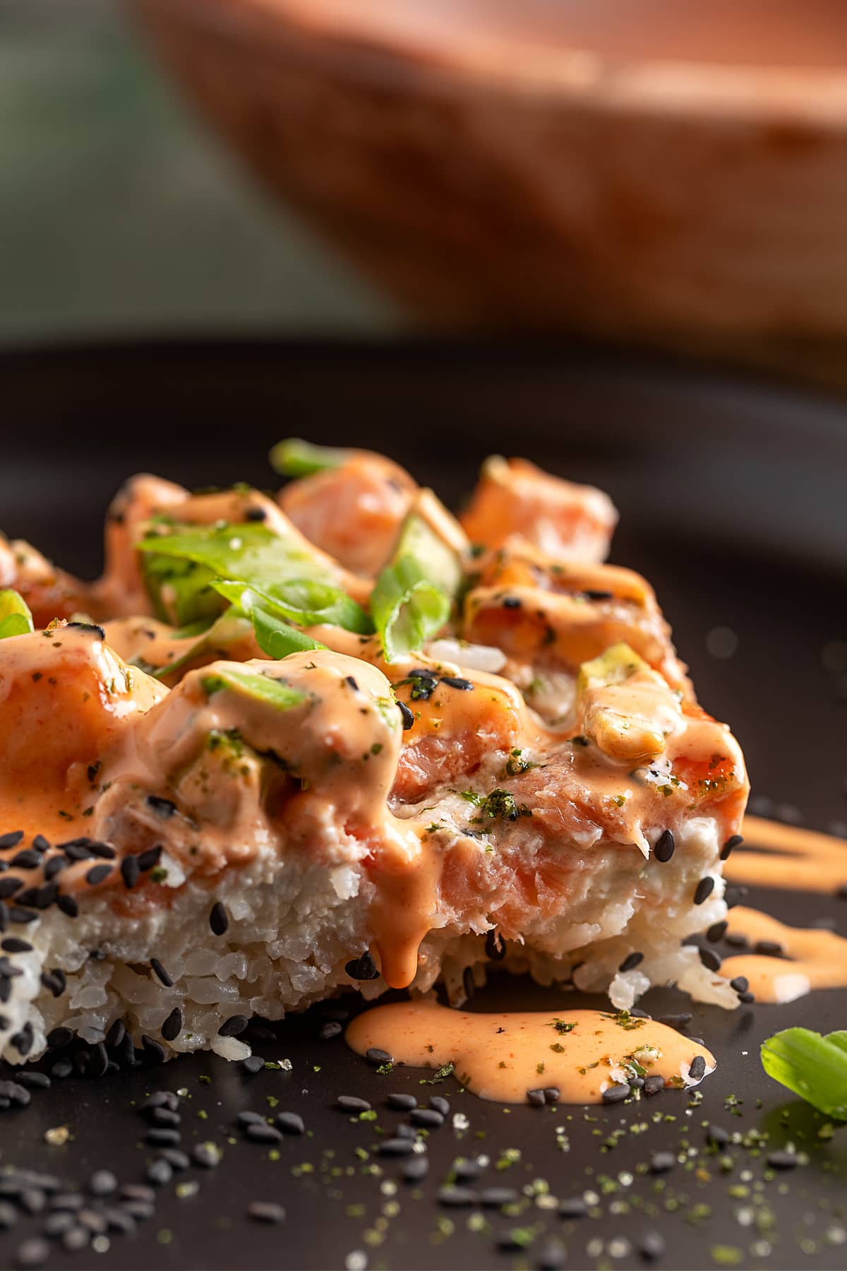 Salmon Crab Sushi Bake served in layered squares with spicy mayo and sesame seeds. Salmon Crab Sushi Bake slice layered with jasmine rice, creamy crab, baked salmon, and spicy mayo on a dark plate