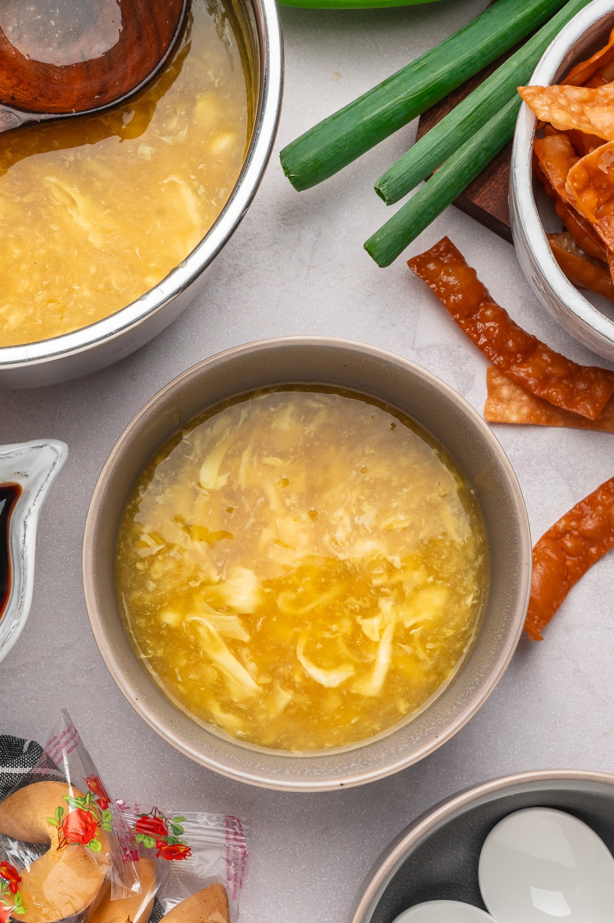 Top-down view of egg drop soup in a bowl showing soft egg ribbons suspended in a lightly thickened golden chicken broth, with wonton strips and green onions nearby Silky egg drop soup with soft egg ribbons in broth