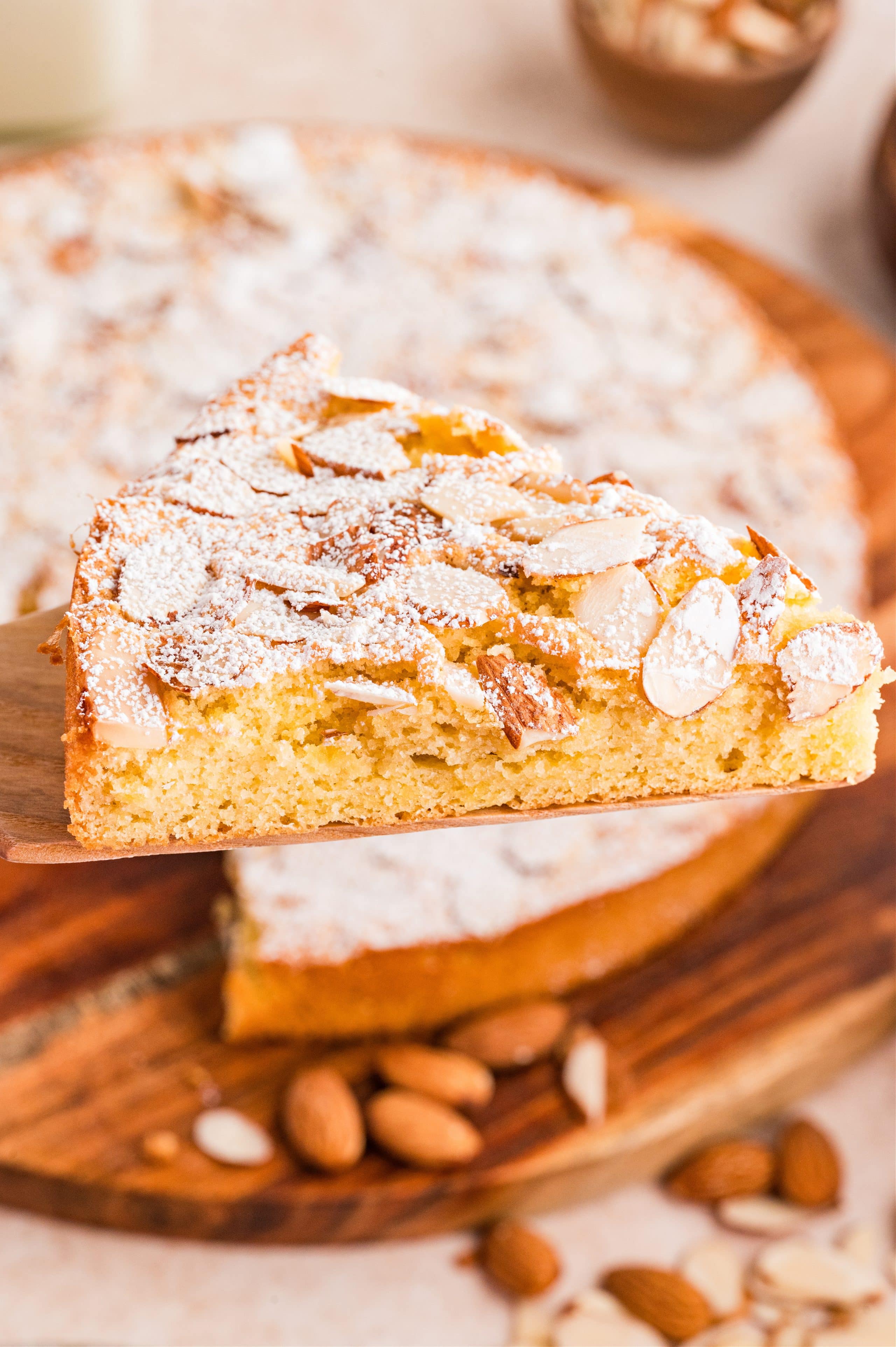 Close-up of a single slice of almond cake topped with sliced almonds and a dusting of powdered sugar on a wooden board. Slice of almond cake with sliced almonds and powdered sugar on top, showing the light, tender crumb