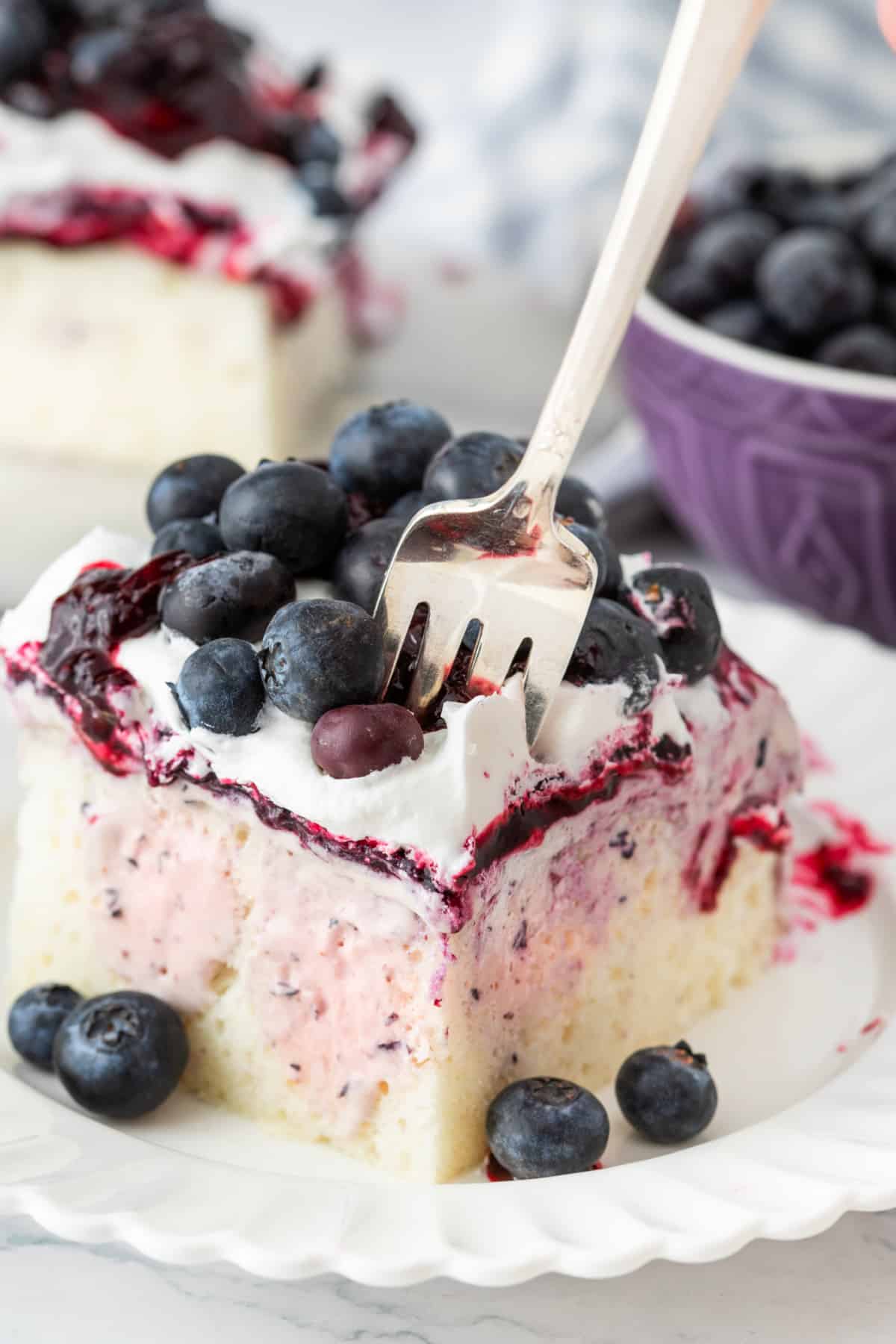 Blueberry cheesecake poke cake bite Fork cutting into blueberry cheesecake poke cake with fresh blueberries and whipped topping