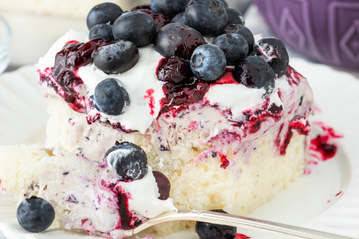 Blueberry poke cake slice with topping Slice of blueberry poke cake topped with whipped topping, blueberry compote, and fresh blueberries