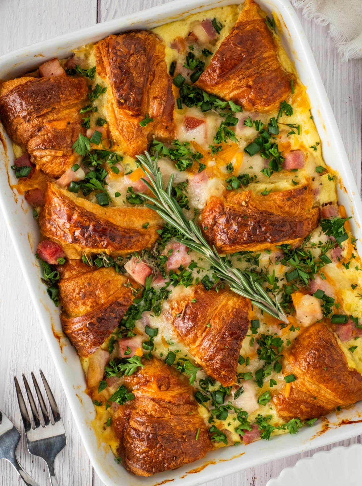 Croissant Breakfast Bake with Ham and Cheese golden croissant breakfast bake with ham, melted Swiss and cheddar cheese, and fresh herbs baked in a white casserole dish