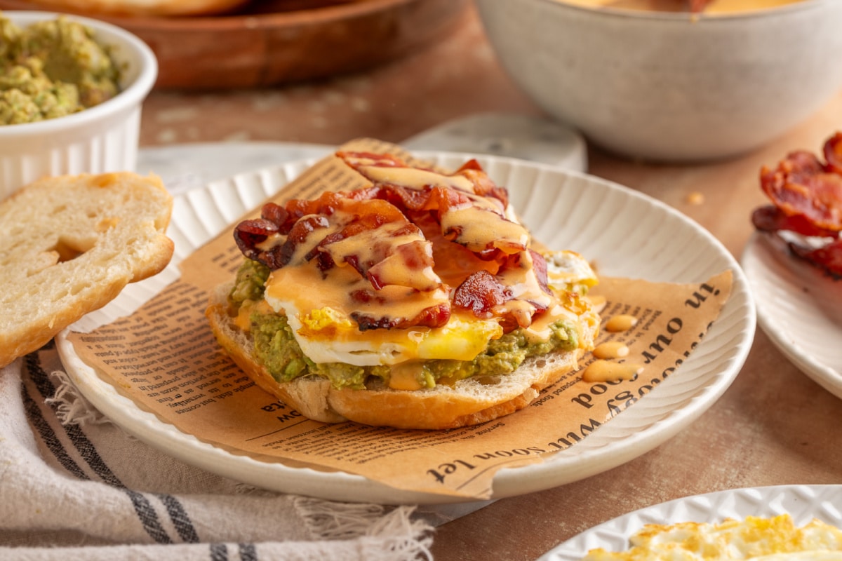 Croissant breakfast sandwich with mashed avocado, sunny side egg, crispy bacon and lime chipotle hollandaise on a white plate