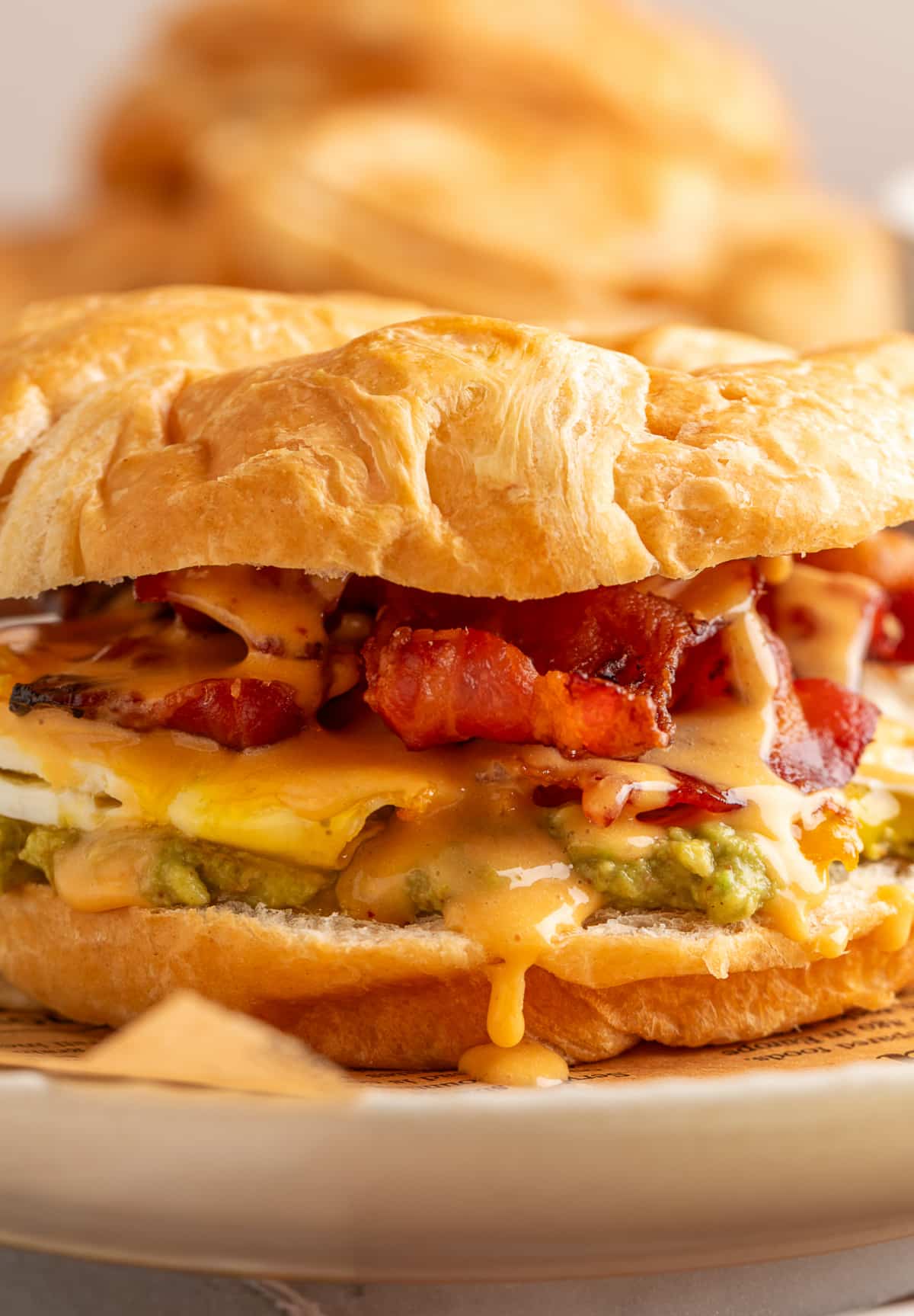 Close up of croissant breakfast sandwich layers with mashed avocado, fried egg, crispy bacon and spicy chipotle hollandaise dripping