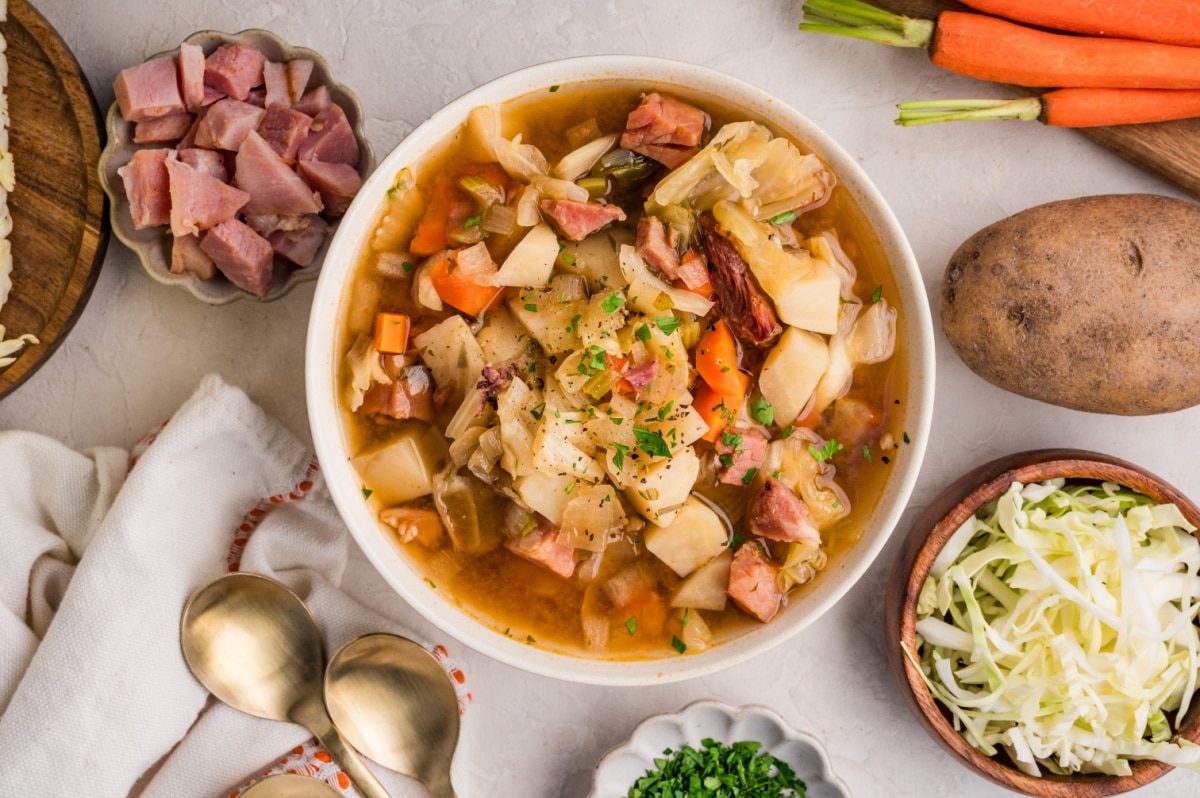 Ham and cabbage soup with ingredients Ham and cabbage soup with potatoes, carrots, cabbage, and ham in a brothy bowl with ingredients around it