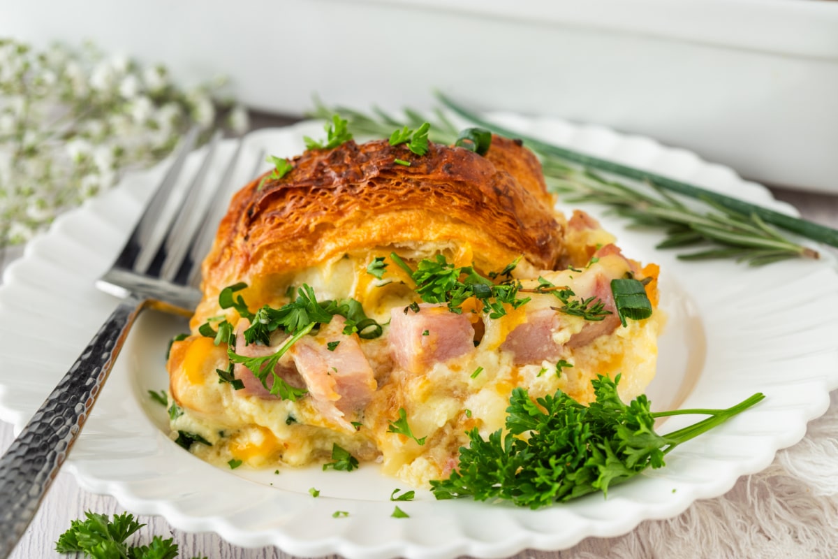 Ham and Cheese Croissant Breakfast Bake Slice slice of ham and cheese croissant breakfast bake served on a white plate with parsley and chives