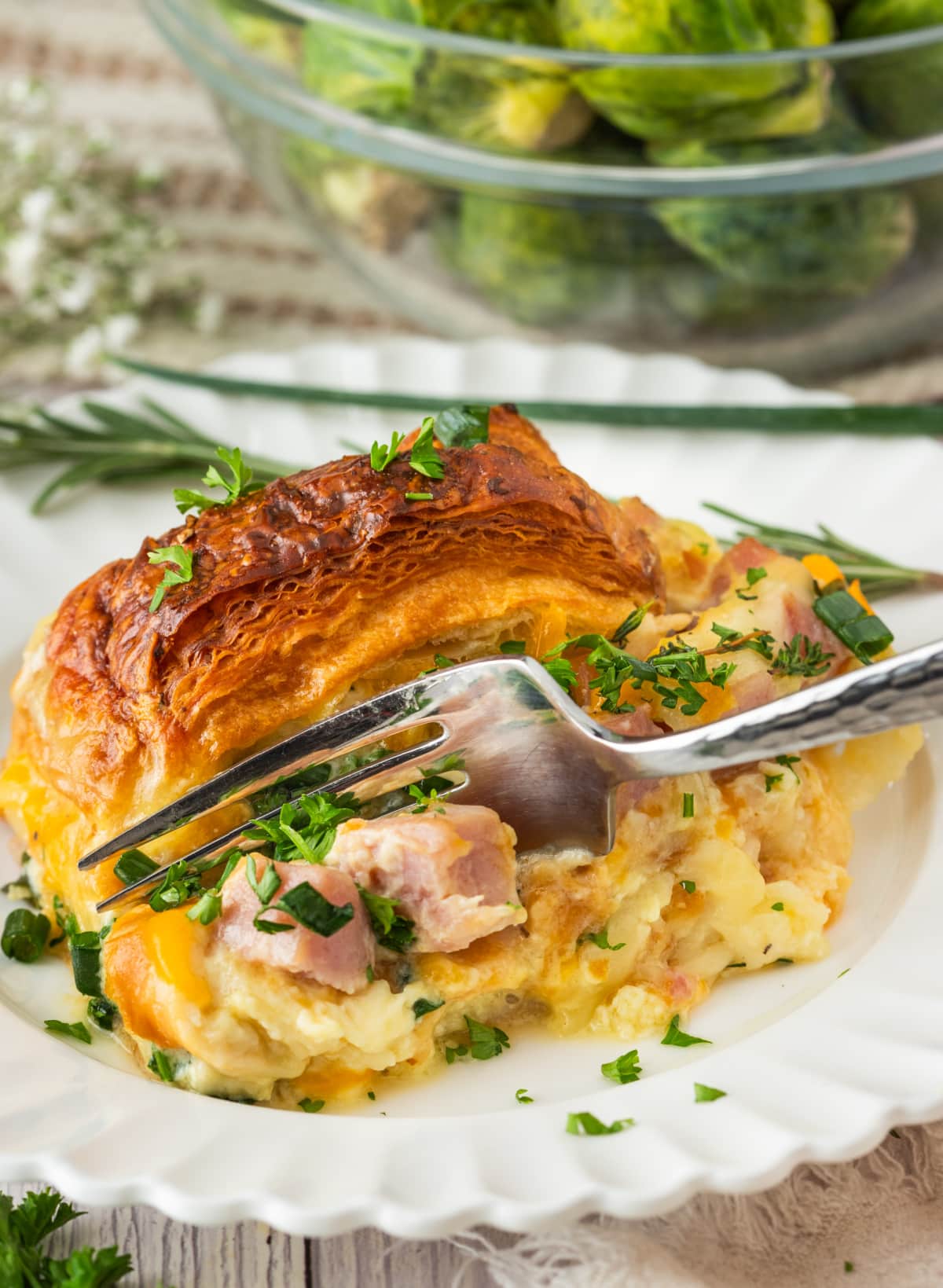 Croissant Breakfast Bake Slice with Ham and Cheese slice of croissant breakfast bake with ham, melted cheese, and egg custard on a white plate with a fork