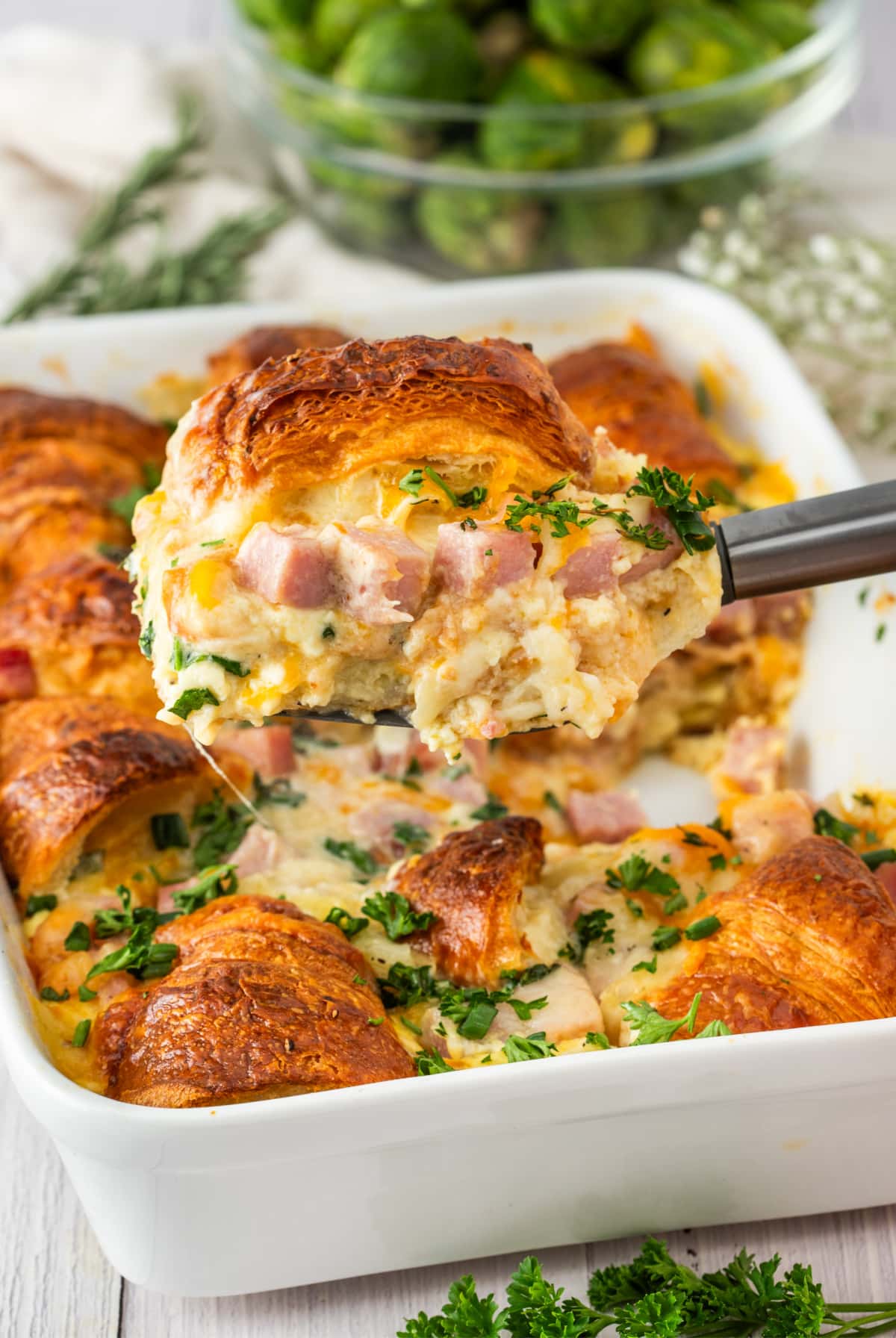 Ham and Cheese Croissant Breakfast Bake Serving ham and cheese croissant breakfast bake served from a white baking dish with golden croissants, melted cheese, diced ham, and fresh herbs