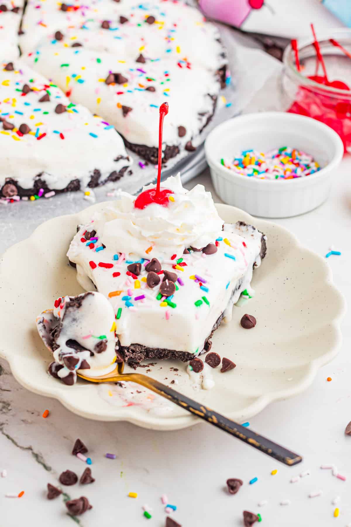 slice of ice cream dessert pizza with Oreo crust topped with whipped cream, cherry, sprinkles, and chocolate chips