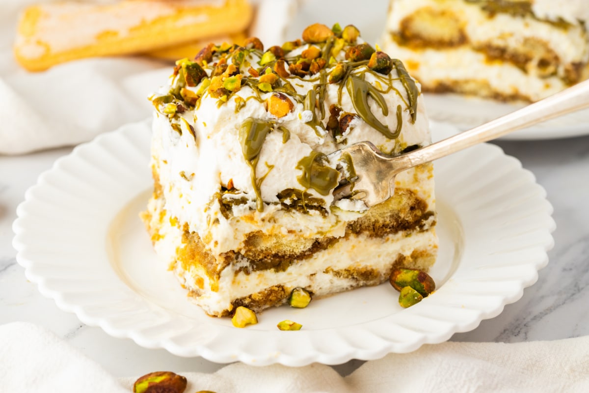 Pistachio Tiramisu Slice with Fork Showing Creamy Layers pistachio tiramisu with fork cutting into layers of mascarpone cream and espresso soaked ladyfingers topped with pistachio drizzle and chopped pistachios