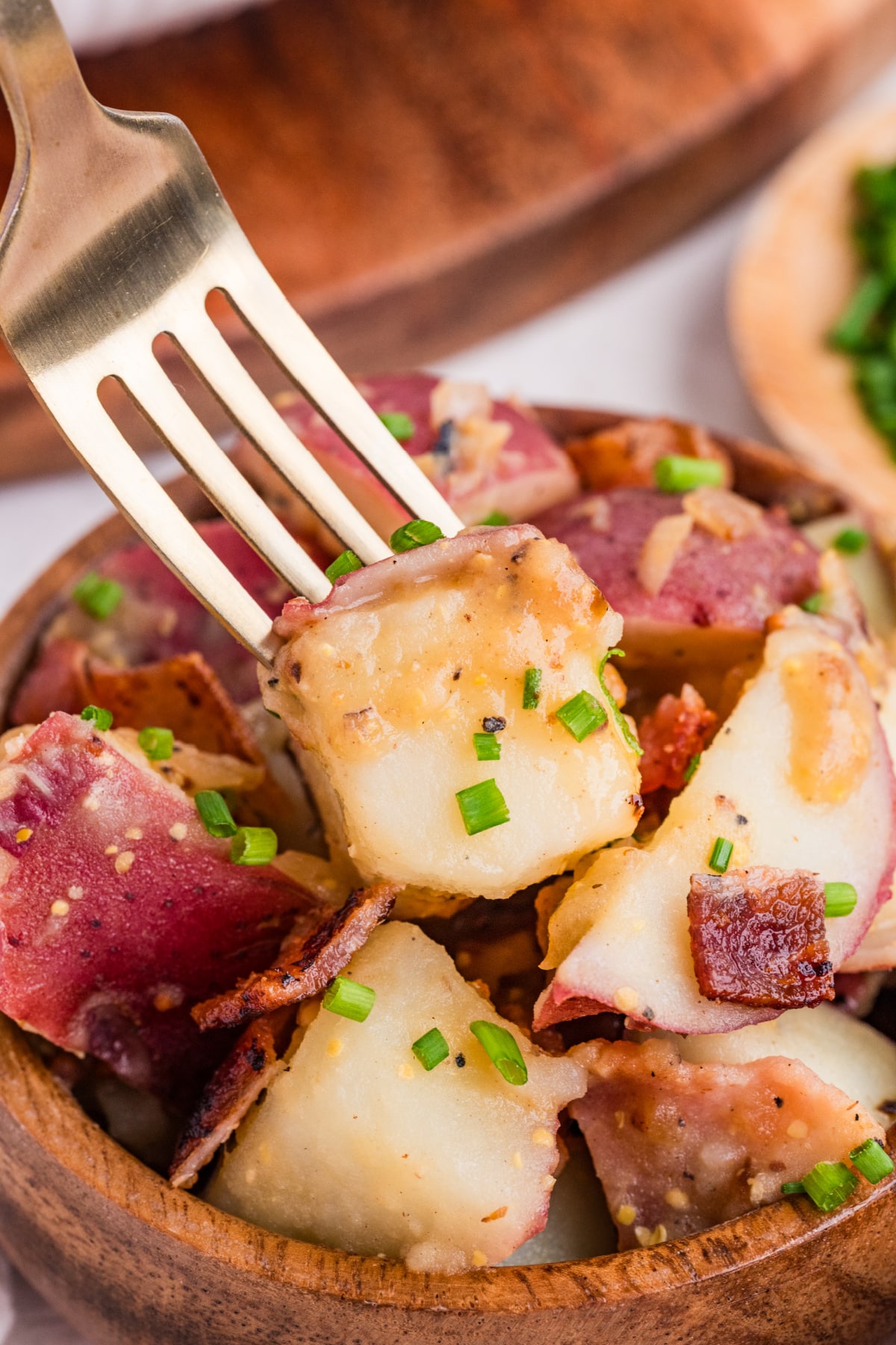 fork bite of potato salad with bacon and mustard dressing fork lifting red potato with bacon, mustard dressing, and chives from warm potato salad
