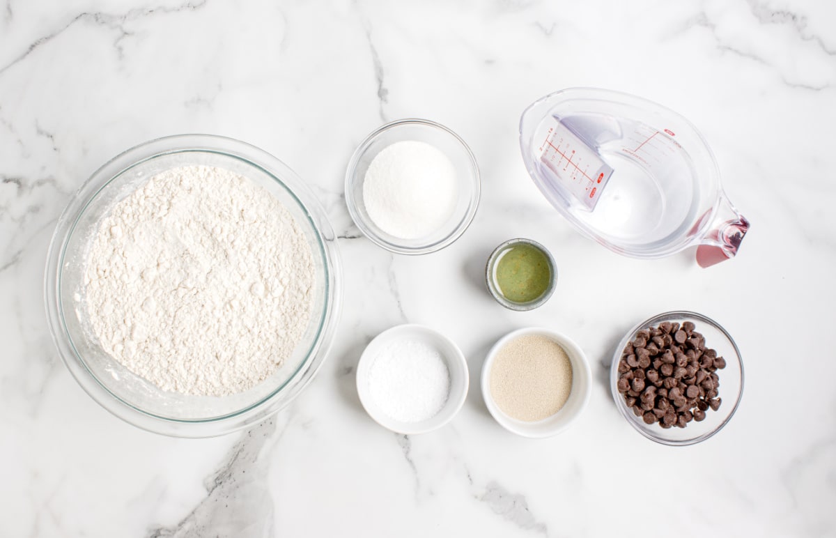 ingredients for chocolate chip bread including flour, yeast, sugar, salt, oil, water, and chocolate chips on a marble surface
