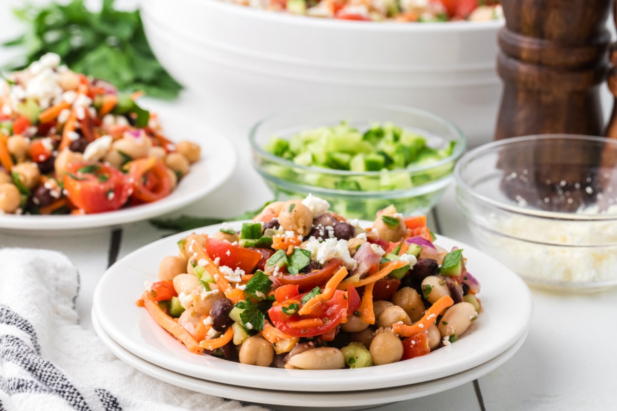Dense bean salad recipe served on a white plate with feta cheese, chickpeas, black beans, tomatoes, cucumber, and parsley